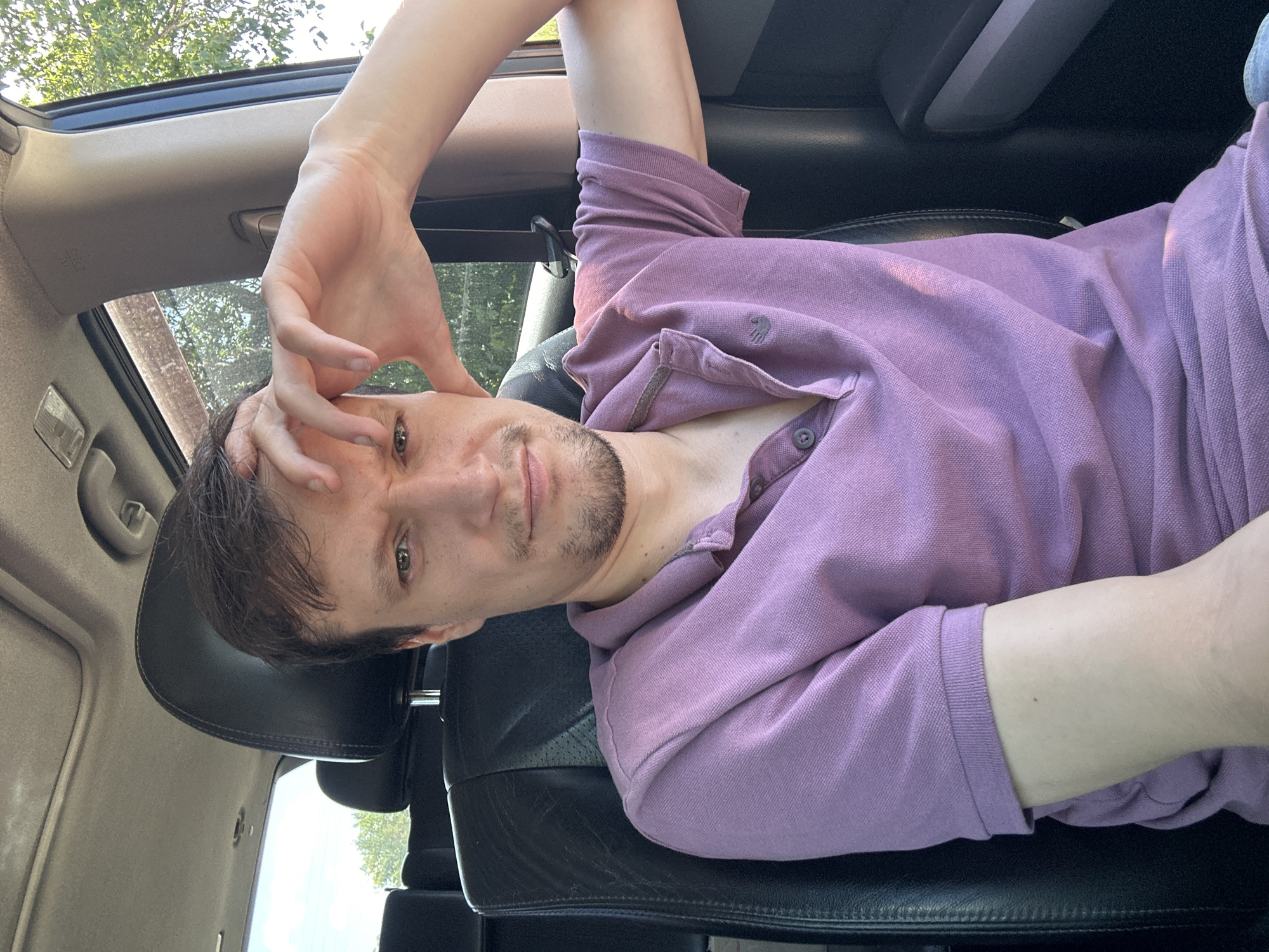 A man is sitting in the driver's seat of a vehicle, likely a car or SUV, with a sunroof visible overhead. The interior of the car appears to be black leather. The man is looking directly at the camera with a slightly furrowed brow and his right hand is raised to his forehead, with his fingers touching his hairline. He is wearing a purple polo shirt with a small logo on the left side of his chest and two buttons at the collar. The man has short brown hair and a short beard. Sunlight is streaming in from the right side of the vehicle, casting a warm glow on his arm and shirt. Outside the car window, trees and foliage are visible, suggesting an outdoor setting, possibly in a rural or suburban area. The specific location in Makiv, Ukraine cannot be determined from the visual information provided in the image.