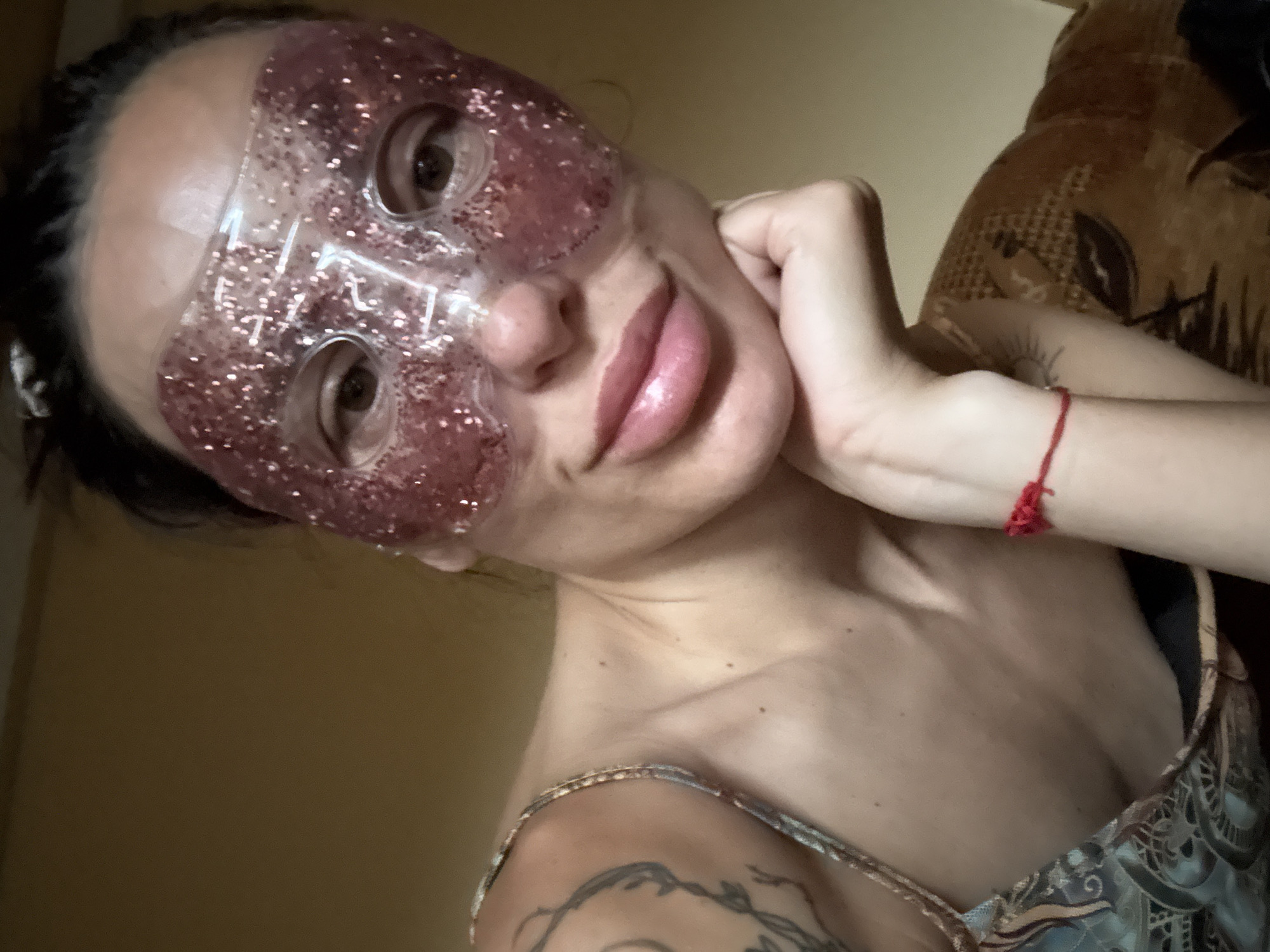 A woman is wearing a pink glitter eye mask and resting her chin on her hand. She has dark hair and is looking at the camera. She is wearing a patterned dress with a strap over her shoulder. She has a tattoo on her arm and a red string bracelet on her wrist. The background appears to be a room with a brown couch. The location is not discernible from the image.