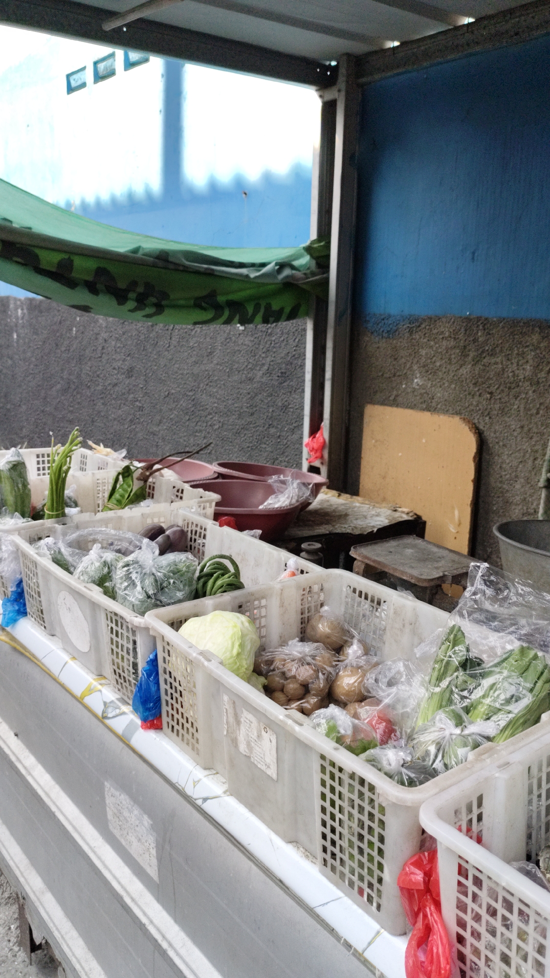 This is a medium shot, eye-level, outdoor photograph of a vegetable stall in Cimahi, Indonesia. The stall is covered by a green tarp with a black camouflage pattern and is set against a rough, grey concrete wall. In the foreground, several white plastic crates are filled with fresh produce. One crate contains long green beans and what appears to be several eggplants. Another crate holds a head of cabbage, potatoes, and other vegetables. A third crate contains green leafy vegetables and what look like some bell peppers. To the left, a green cucumber and a bundle of green onions are visible. In the background, there are two large, maroon-colored bowls, one on top of the other. A wooden board leans against the grey wall. The lighting is diffused, suggesting an overcast day or the stall being in a shaded area.