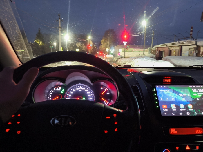 The image depicts the interior of a vehicle, specifically from the driver's perspective, stopped at an intersection in Sarapul, Russia, during twilight or nighttime conditions.
A human left hand is gripping the black multi-function steering wheel, which features illuminated red buttons on the left spoke and a central "KIA" logo. The dashboard gauges are illuminated in red and white, displaying a tachometer (left), speedometer (center, in km/h), fuel level, and engine temperature. The tachometer indicates the engine is idling. A large aftermarket infotainment system is mounted in the center console, displaying "19:33" as the time, "98.10" as a radio frequency, and various application icons. Above this, a smaller digital clock shows "17:32". Below the infotainment system, a climate control display indicates an interior temperature setting of "20.0" degrees Celsius.
Through the dirty or condensation-covered windshield, the exterior environment shows a dark blue sky. Multiple streetlights are active, illuminating the scene. A prominent traffic light ahead displays a red signal, indicating the vehicle is stopped. Additional red and faint green traffic signals are visible further down the road. The road surface appears to be asphalt with accumulated snow and slush on the shoulders and median, confirming winter conditions. Utility poles with overhead wires line the street. On the right side of the road, several low-rise buildings, possibly garages or commercial structures, are visible, with a distinctive red-and-white striped chimney-like structure behind one. Distant vehicle lights are also discernible.
