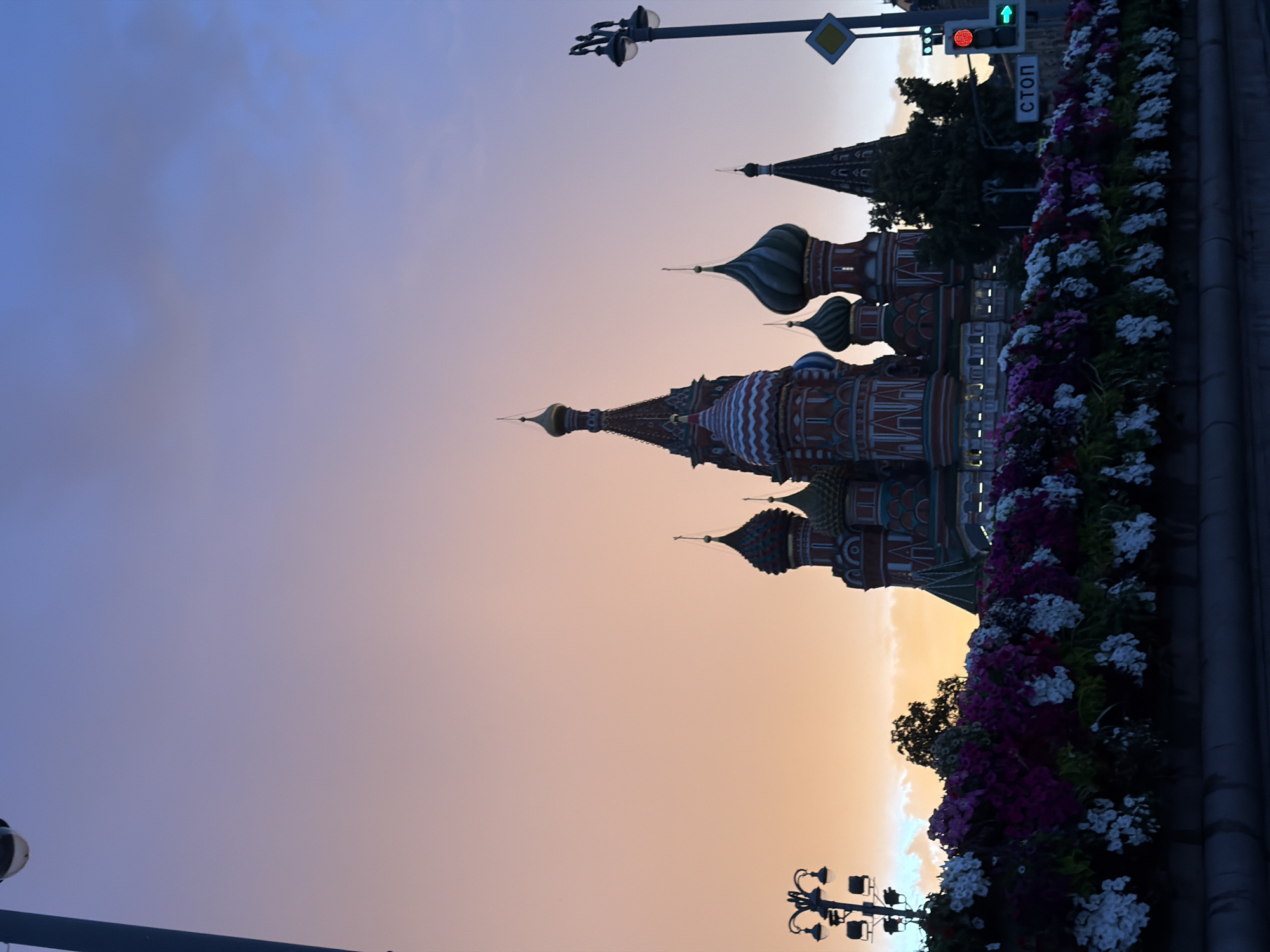 The image shows Saint Basil's Cathedral in Moscow, Russia, during what appears to be either sunrise or sunset, given the gradient of colors in the sky, ranging from soft blues and purples to warm oranges and yellows. The iconic onion domes of the cathedral are silhouetted against the colorful sky, with intricate architectural details visible. In the foreground, a flower bed overflows with a vibrant display of purple and white flowers, adding a touch of natural beauty to the scene. A street lamp with multiple lights stands to the left, and traffic lights and a diamond-shaped road sign are visible above the flowers to the right. The word "СТОП" (STOP in Russian) is visible on a street sign. The overall mood is serene and picturesque, capturing a moment of quiet beauty in an urban setting. The image is oriented vertically.