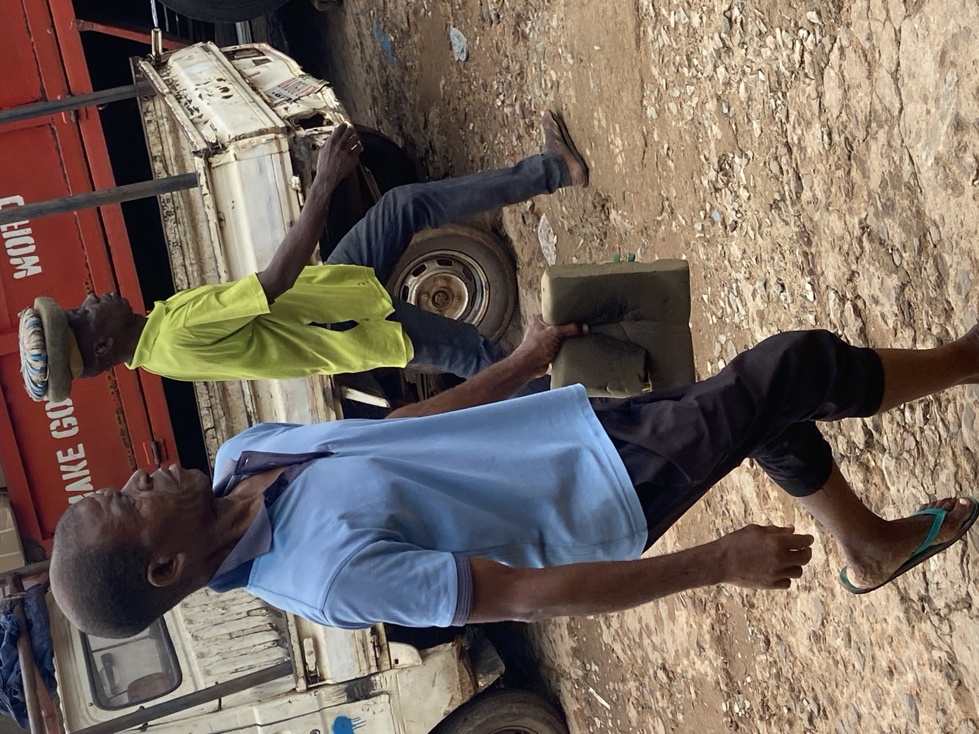 In Ogbunike, Nigeria, two men are walking in a dirt-covered area next to a truck. The man in the foreground, wearing a light blue polo shirt and dark pants, is carrying a green cushion. He is walking towards the left of the frame, with his head turned to the right. The second man, in a neon yellow shirt and dark pants, is walking behind him, carrying a cushion on his head. The truck is white with red accents and appears to be old and worn. The ground is uneven and rocky, with some loose gravel. The overall scene suggests a busy, working environment.