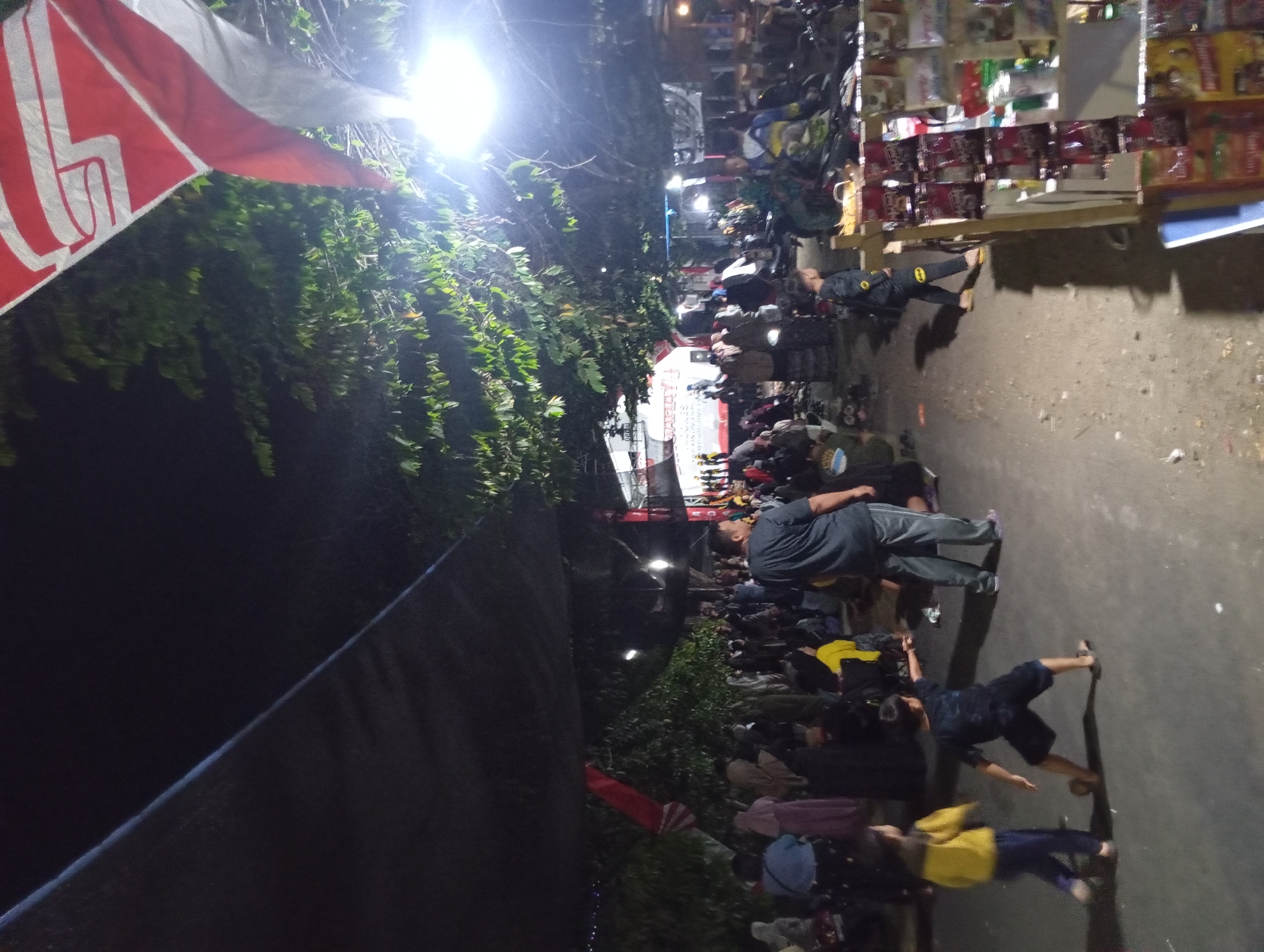 In Jogosari, Indonesia, a nighttime outdoor gathering is depicted. A crowd of people is present, with some standing and others seated. The scene is illuminated by a bright overhead light, casting shadows and highlighting a large black tarp stretched overhead. Trees with dense foliage are visible on the right side of the image, with a flag featuring red and white bands prominently displayed in the upper right corner. A small stand with various packaged items, possibly snacks or convenience goods, is set up on the right. In the foreground, two young boys are interacting, with one wearing a yellow jacket and blue pants, and the other in a dark blue outfit. The ground is unpaved.