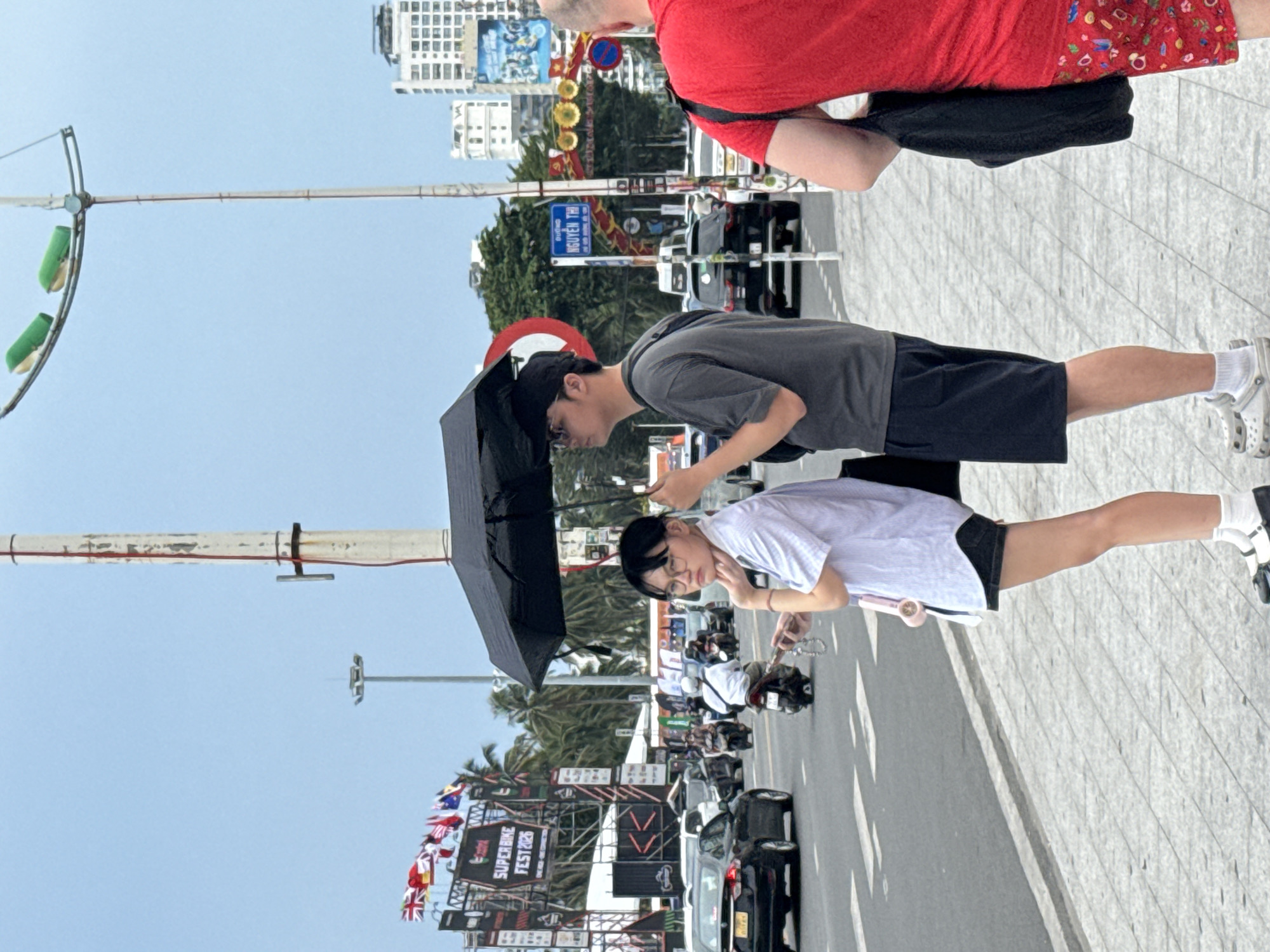 A photo shows an outdoor scene in Nha Trang Ward, Vietnam, with pedestrians on a paved area alongside a street with traffic.
A person in a gray shirt and black shorts holds a black umbrella over themselves and another individual. The second person, wearing a white shirt and black shorts, stands beside them. The two seem to be engaging with a phone or camera.
On the right of the frame, a man in a red shirt with colorful shorts is partly visible.
The street has vehicles, including motorcycles and cars. In the background, there are buildings, trees, and signs, including one that reads "NGUYEN THI." A large banner promoting a "SUPER BIKE FEST 2026" with various flags is also visible.
The weather appears to be sunny.