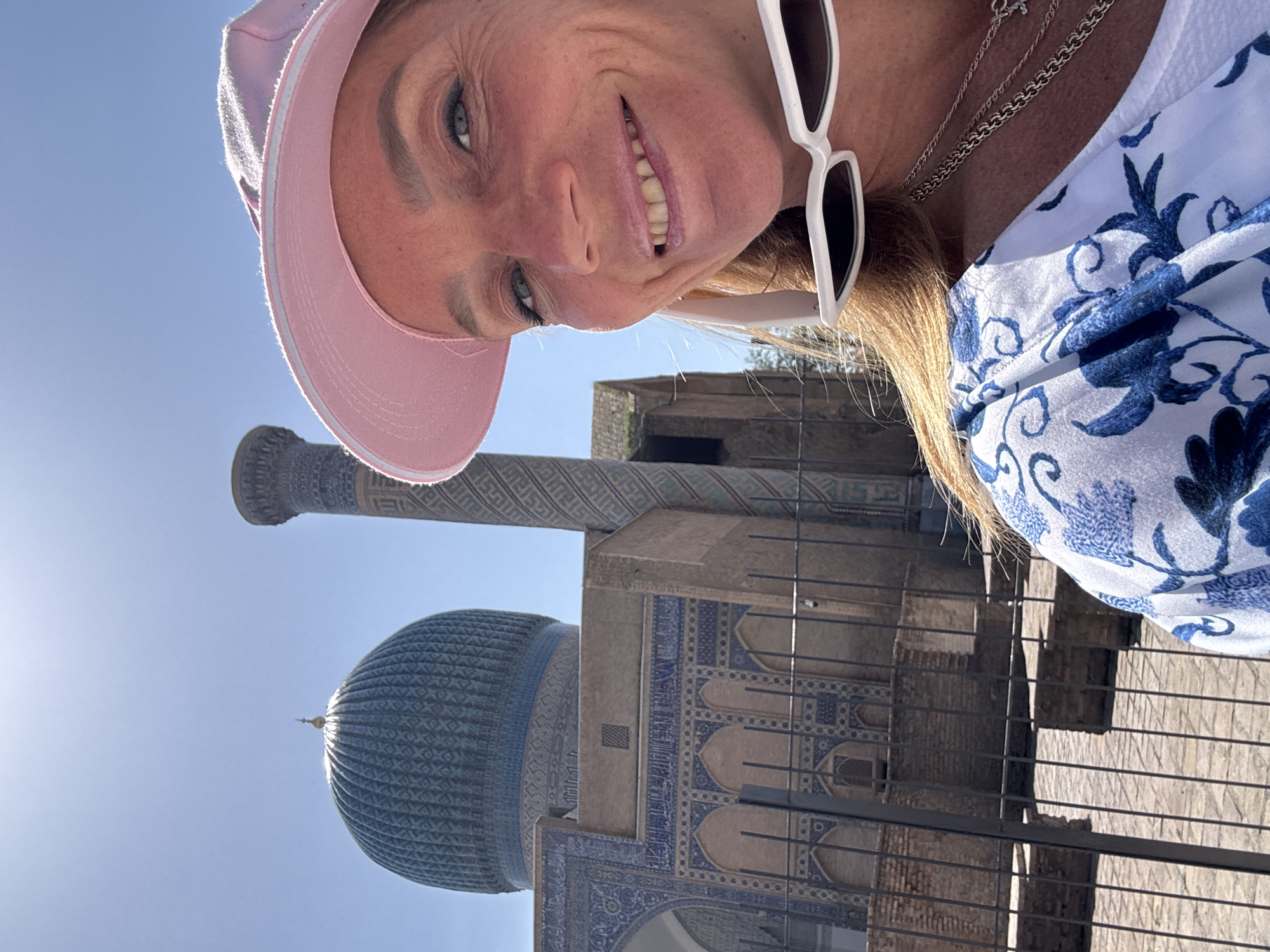 A selfie-style photo shows a woman with a pink hat and white sunglasses standing in front of an architectural monument in Samarkand City, Uzbekistan. The woman has blonde hair, blue eyes, and is smiling at the camera. She is wearing a white and blue patterned top with silver necklaces. Behind the woman, there is a large building with a blue dome and intricate tilework. A tall minaret is visible to the left of the building. The sky is clear and blue. A metal fence is in the foreground, partially obscuring the view of the building. The photo has a slight tilt.