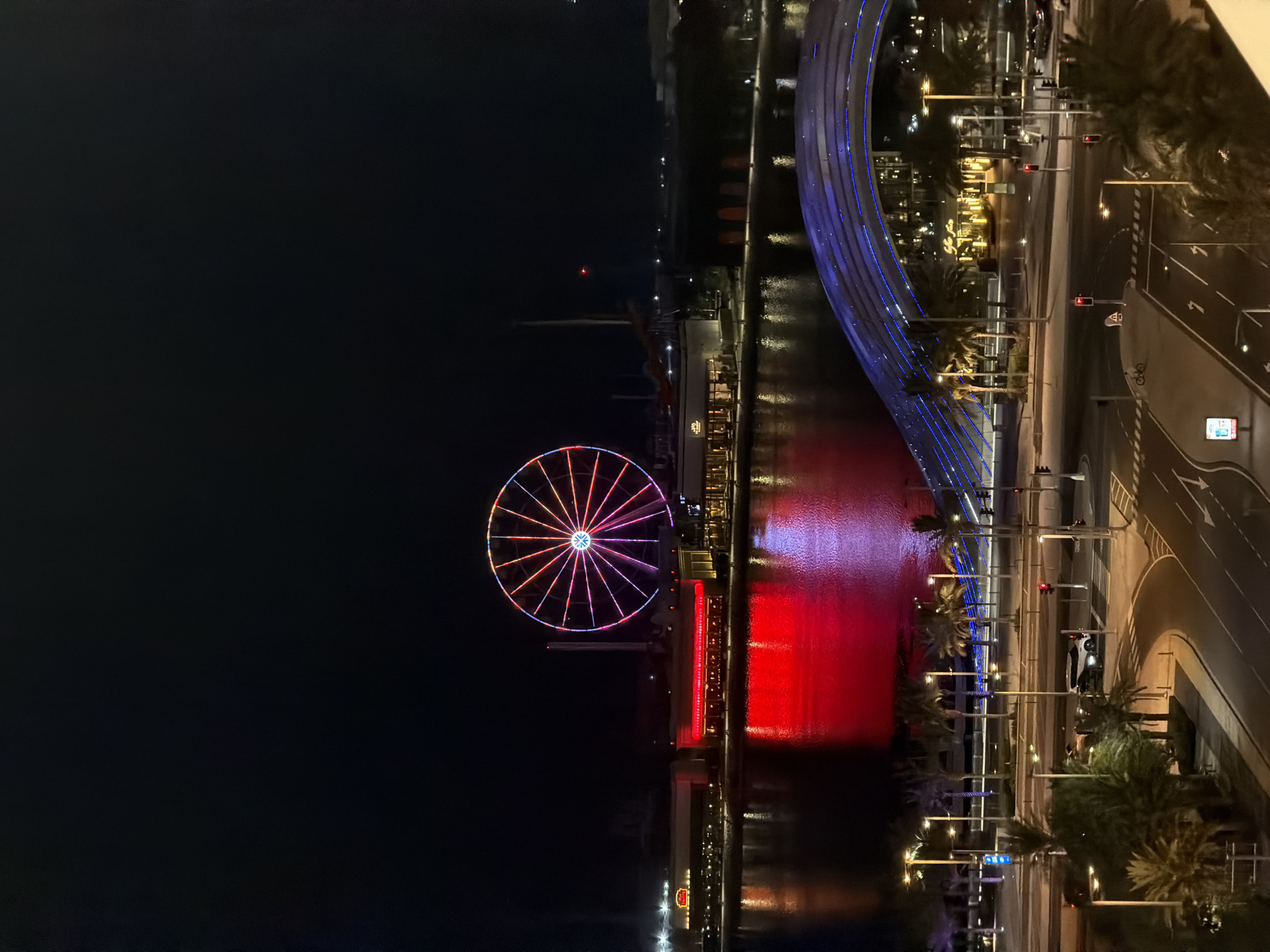 An elevated, wide-angle nighttime shot captures a Ferris wheel and illuminated buildings reflected in a body of water, with a bridge curving over a street in the foreground. The Ferris wheel, a prominent feature, is lit with a colorful pattern of red, orange, pink, and purple lights against the dark sky. Below it, a strip of buildings glows with warm yellow and red lights, their reflections shimmering on the dark water. A pedestrian bridge with a blue light-up design arches over a street below, which is sparsely populated with a few cars and palm trees lining the sidewalk. The scene is set in an urban environment at night, with the lighting suggesting a lively entertainment district.