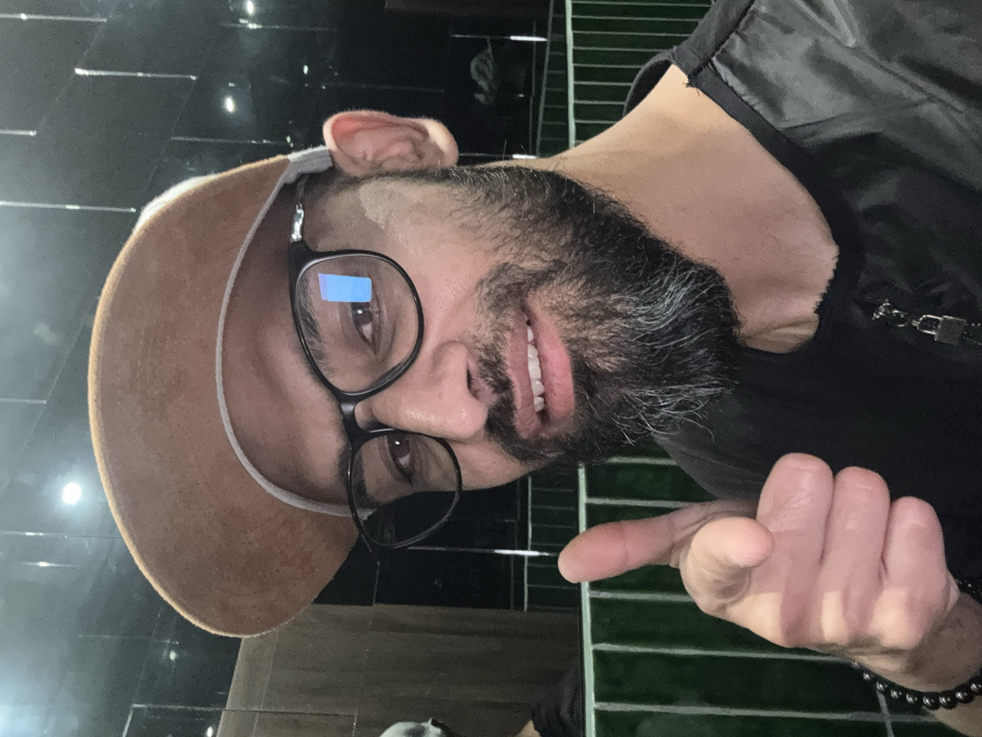 A selfie features a man with a beard and glasses, wearing a tan baseball cap and a black jacket. He is pointing at the camera with his right hand. The background is dark, with some reflections of lights and what appears to be green tiled walls. The scene is not identifiable as being in Plovdiv, Bulgaria.