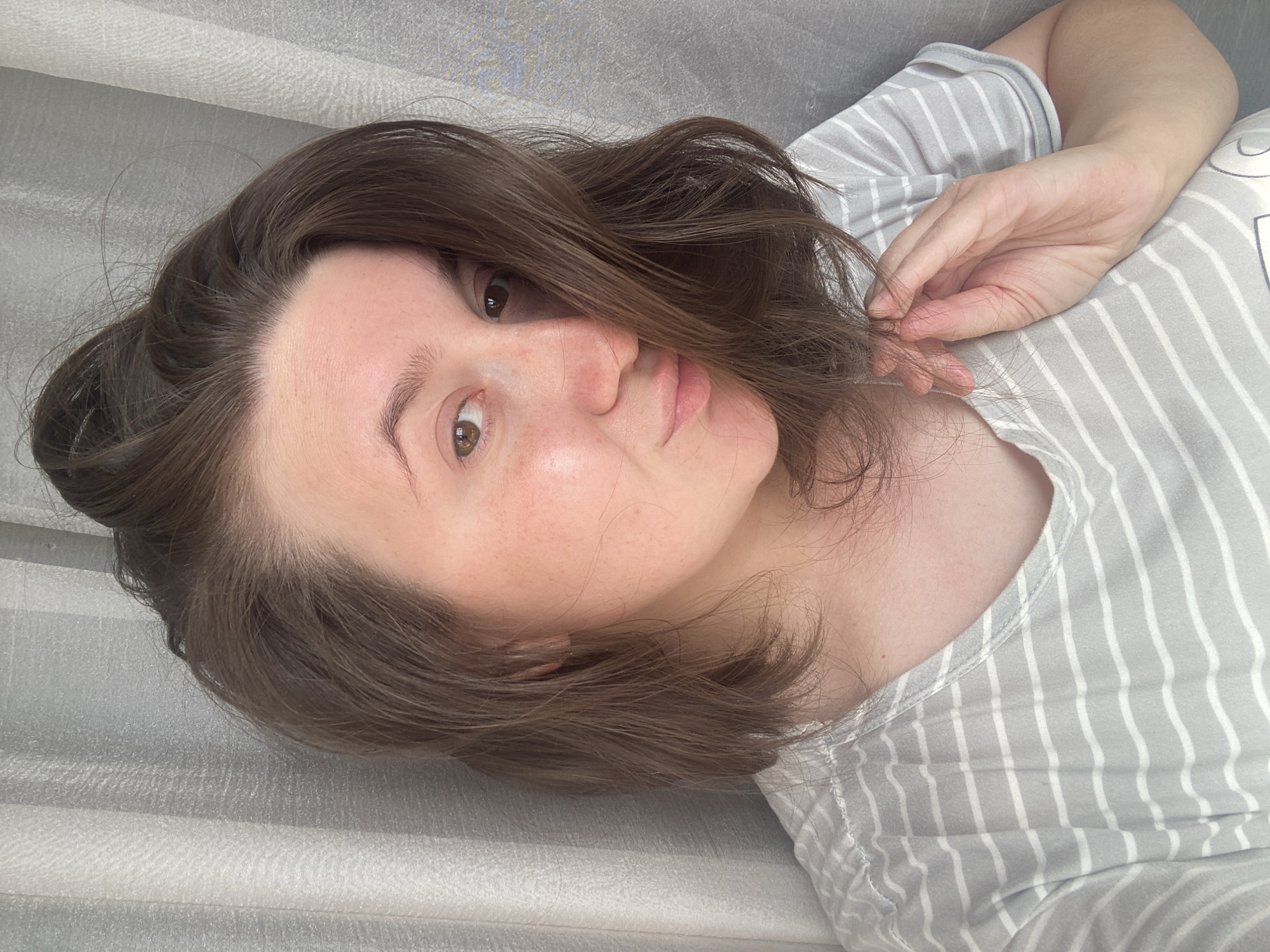 A young woman is depicted in a close-up selfie, taken indoors in Chaikovsky, Russia. The image is rotated 90 degrees, showing her lying down with her head to the left side of the frame. She has medium brown, wavy hair that falls around her face, and her eyes are a warm brown. Her fair skin shows a scattering of light freckles across her nose and cheeks.
She looks directly at the camera with a soft, calm expression, her lips forming a subtle pout or slight smile. She is wearing a light grey top with thin white vertical stripes. One hand is visible near her face, with her fingers gently touching strands of her hair. The background is a soft, light-colored textured fabric, likely a curtain or bed sheet, contributing to a diffused and intimate lighting environment. The soft, natural light suggests it is daytime. No text is visible in the image. The overall mood is relaxed and serene.