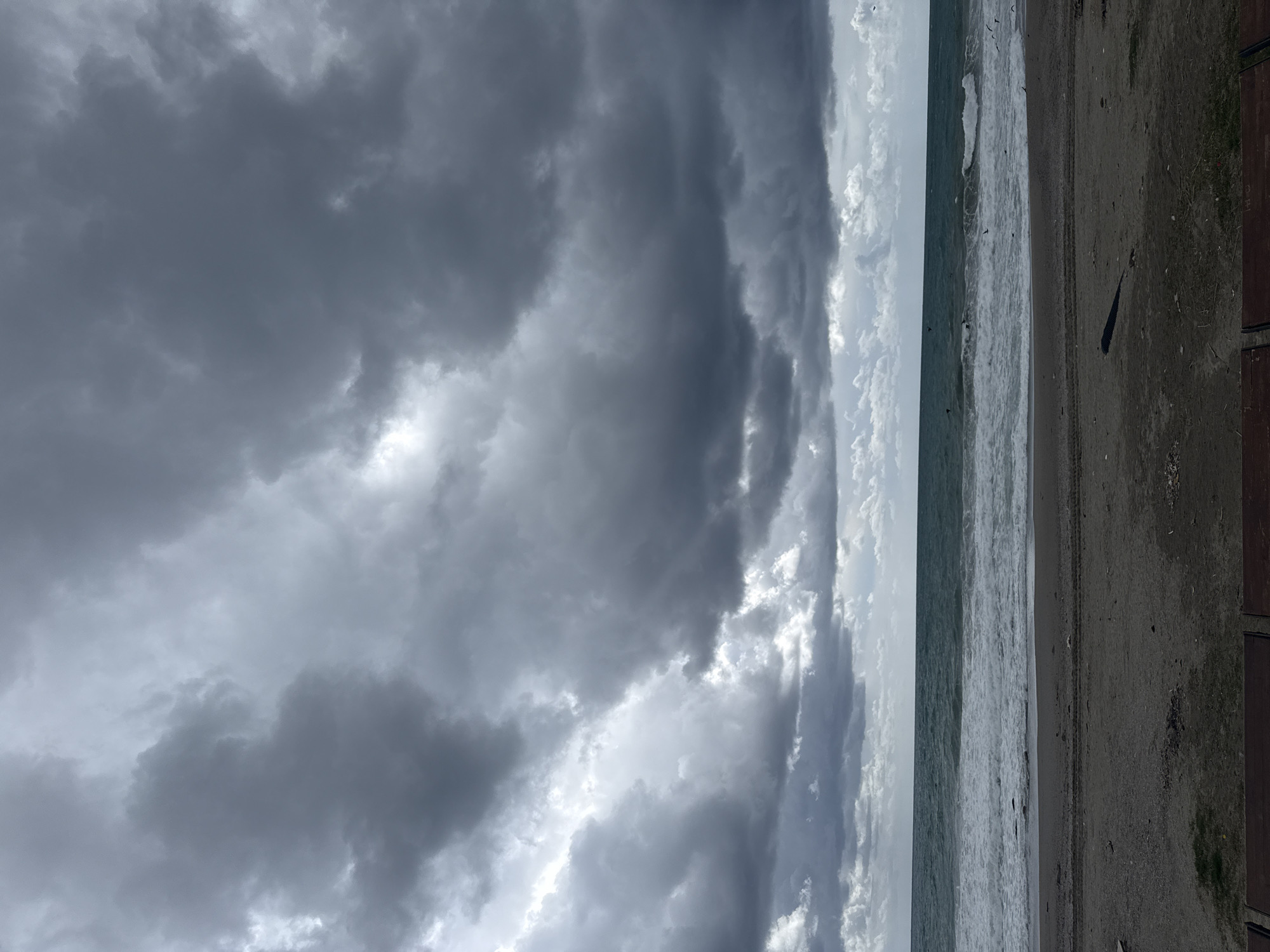 The image shows a beach scene in Alanya, Turkey, under a heavily clouded sky. The sea is visible, with waves breaking near the shoreline. The beach itself is sandy and appears to be slightly wet. To the right of the frame, a wooden structure is partially visible, possibly a walkway or part of a building along the coast. The cloud cover dominates the scene, ranging from dark, heavy clouds to lighter, more scattered formations near the horizon. The overall atmosphere is overcast and possibly stormy.