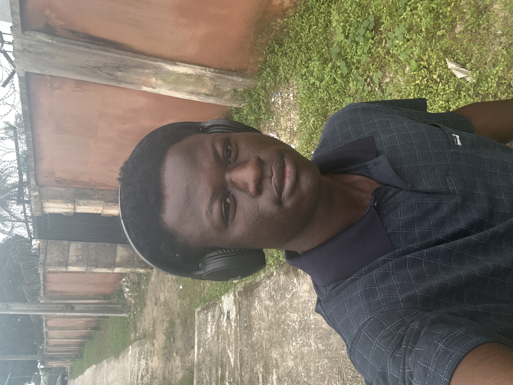 A medium-shot, selfie-style photo shows a young Black male wearing black over-ear headphones and a dark blue collared shirt with a subtle checkered pattern. He is looking directly at the camera with a neutral expression. The background consists of an outdoor scene with a concrete walkway to the left and a reddish-brown wall to the right. Patches of green grass and some debris are visible between the walkway and the wall. The location is identified as Badagry, Nigeria, suggesting an urban or suburban environment typical of the region. The lighting is natural, indicating it was taken during the day.