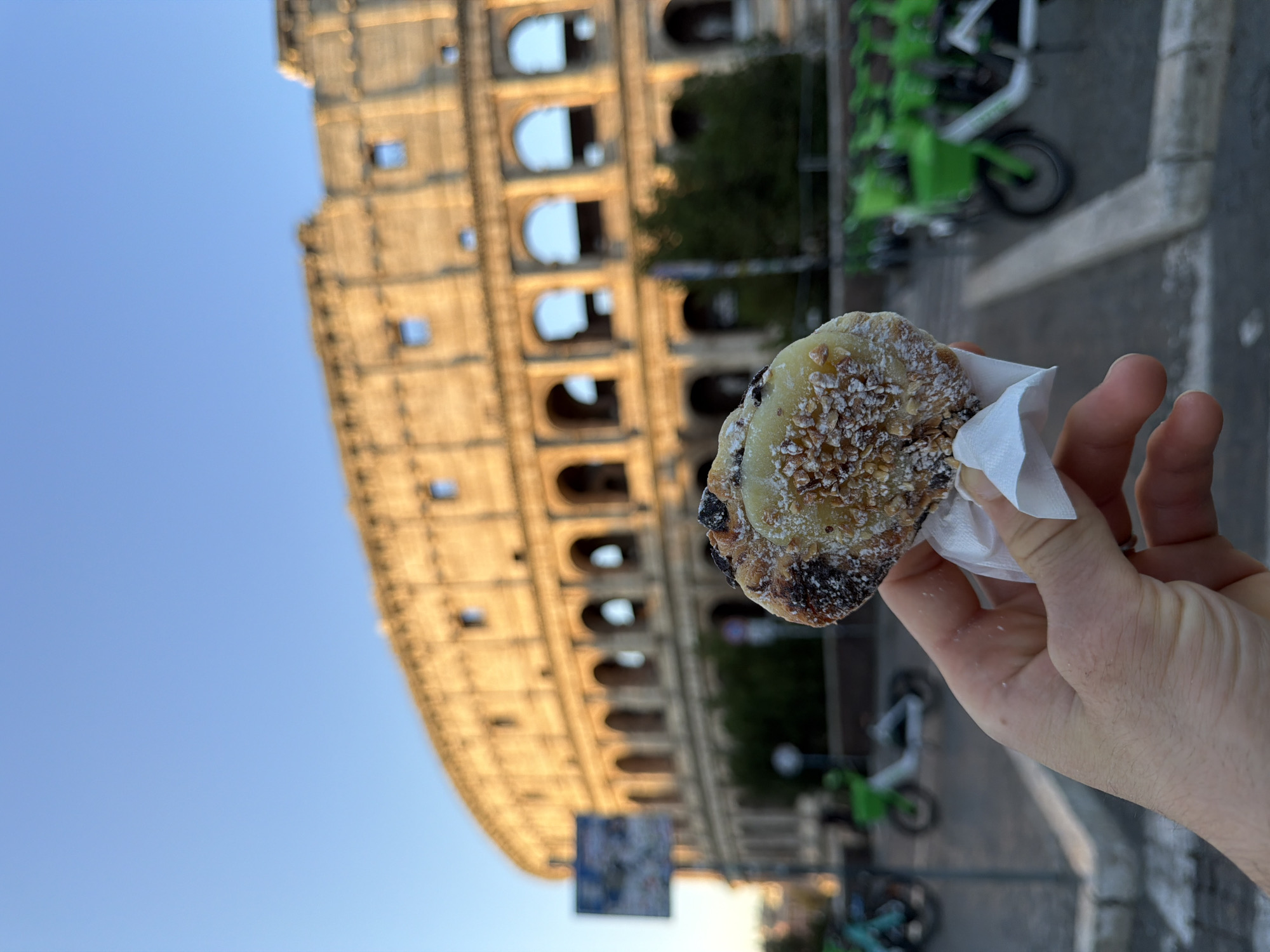 A person's hand holds a pastry in front of the Colosseum in Rome, Italy. The pastry is covered in a light-colored topping and chopped nuts, with visible dark chocolate pieces. The Colosseum, bathed in warm sunlight, is in the background, with its iconic arches and weathered stone. In the middle ground, a street sign and several bright green electric scooters are parked. The sky above is a clear, pale blue.