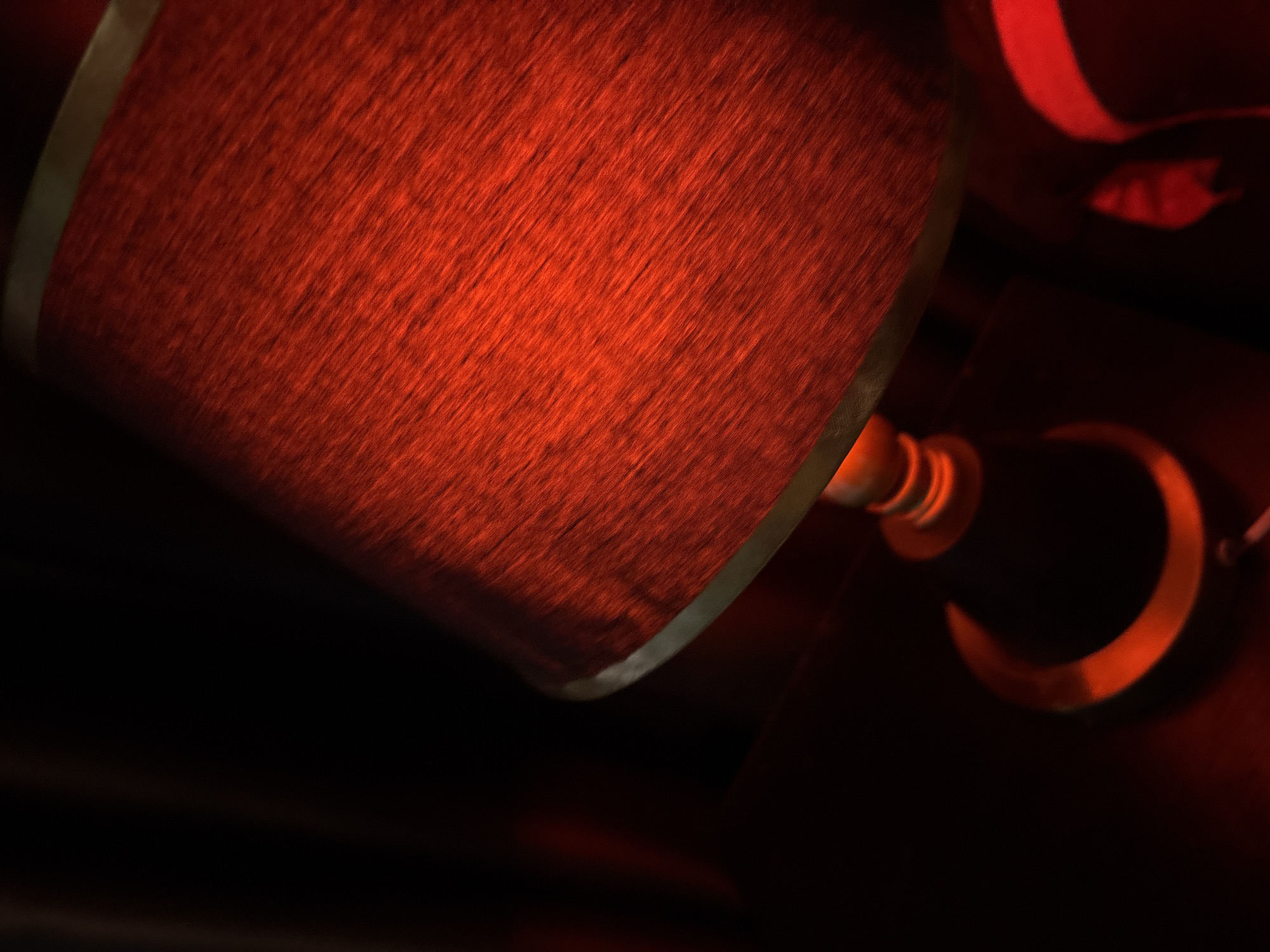 An upward-angled, dimly lit photograph captures a red lampshade. The lampshade is textured, with visible fibers running diagonally. A thin, metallic band circles the top and bottom of the lampshade. The base of the lamp, made of a darker material with metallic accents, is visible below. The surrounding environment is dark, with hints of red illumination reflecting off surfaces. The overall impression is that of a cozy, ambient setting, possibly within a bedroom or living room in a home in Obosi, Nigeria. There are no people or visible actions in the frame.