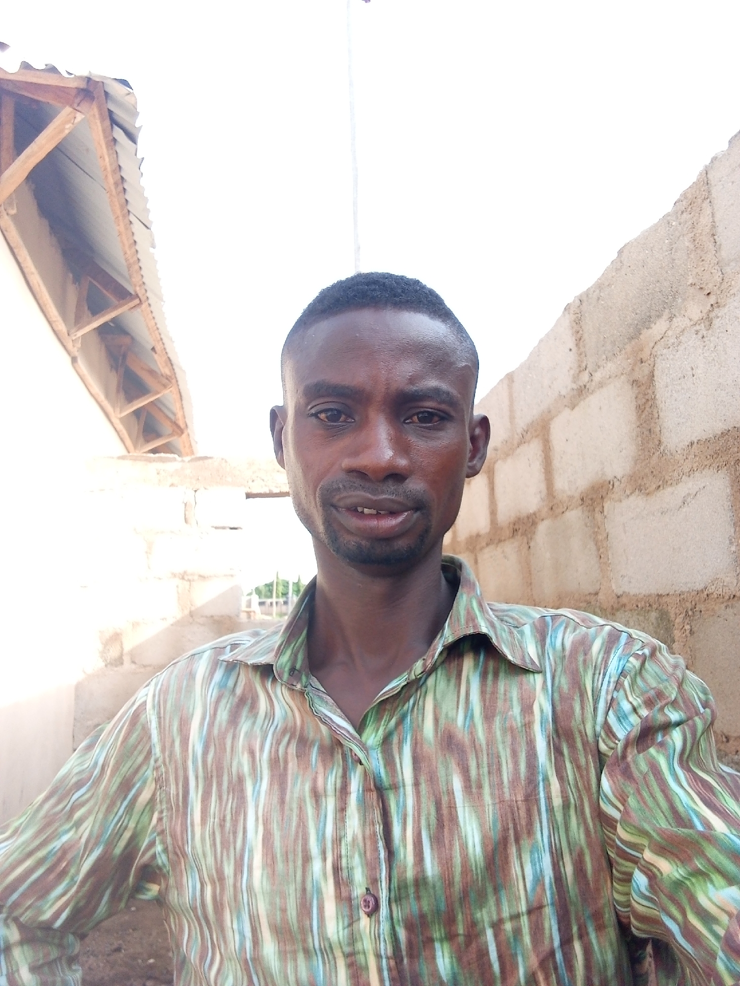 A man of African descent, likely in his 30s or 40s, is centered in a portrait-style photograph. He has short, dark hair, a light mustache, and a short beard, with a neutral expression. He is wearing a long-sleeved button-up shirt patterned with vertical stripes in shades of brown, green, and light blue.
The scene is outdoors, with the man standing against a background that suggests a simple, possibly unfinished, setting. To his right, a rough, unpainted cinder block wall is visible. To his left, there's a light-colored wall and an exposed wooden roof structure topped with corrugated metal sheeting, indicating a basic building or shed.
The photograph appears to have been taken during the day under bright, natural sunlight, evidenced by strong highlights and shadows, particularly on the left side of the image. The man is looking directly into the camera. There is no visible text in the image. This setting, characterized by cinder block walls and corrugated roofing, is consistent with construction often found in places like Adonkolo, Nigeria.
