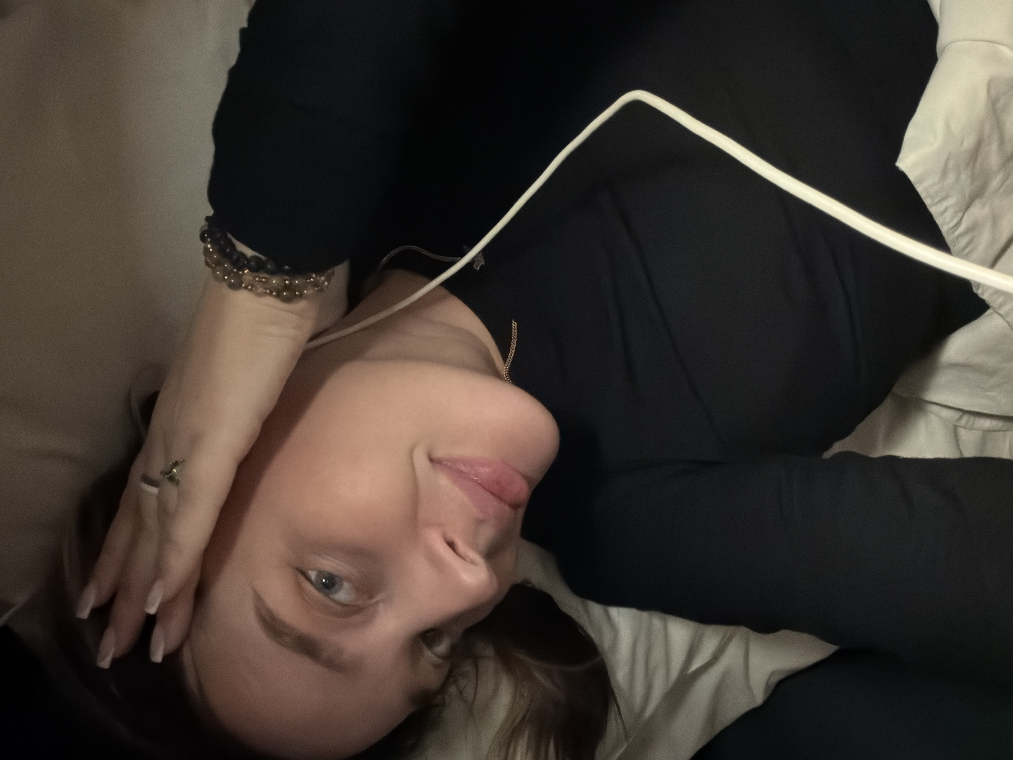 The image shows a young woman lying on her back, her head slightly tilted, gazing directly towards the viewer with a calm expression and a subtle smile. Her blue eyes are prominent, and her dark brown hair is partially visible around her head. She is dressed in a black long-sleeved top or dress. Her right hand, adorned with a ring on her ring finger and manicured nails, rests gently near her head. On her left wrist, she wears multiple beaded bracelets, some dark and others with a golden sheen. A thin gold-colored chain necklace is visible around her neck. A white cable, likely a charging or earphone cable, extends across her chest and shoulder. She is situated indoors, appearing to be on a bed or sofa, indicated by the rumpled white or light-colored fabric surrounding her. The ambient lighting is dim, suggesting a low-light environment or nighttime. There are no discernible specific location cues beyond an indoor setting, nor is any text visible.