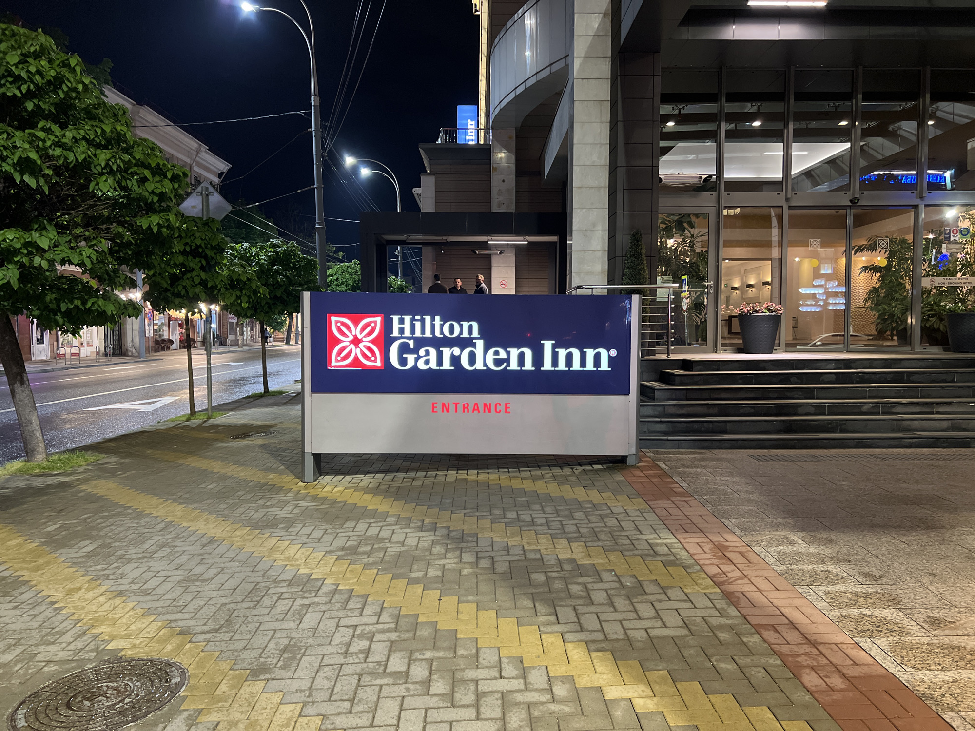 The image shows a Hilton Garden Inn hotel at night. A large sign stands on the sidewalk in front of the hotel, illuminated with the hotel's name and logo, and the word "ENTRANCE" in red. The sidewalk is paved with interlocking bricks in gray and yellow patterns. A street with sparse traffic and streetlights is visible to the left, lined with trees and buildings. Two people are visible standing under a small awning at the entrance to the building, engaged in what looks like smoking. The entrance to the hotel is visible behind the sign, with glass doors and plants inside. The image was taken in the city of Krasnodar, Russia.