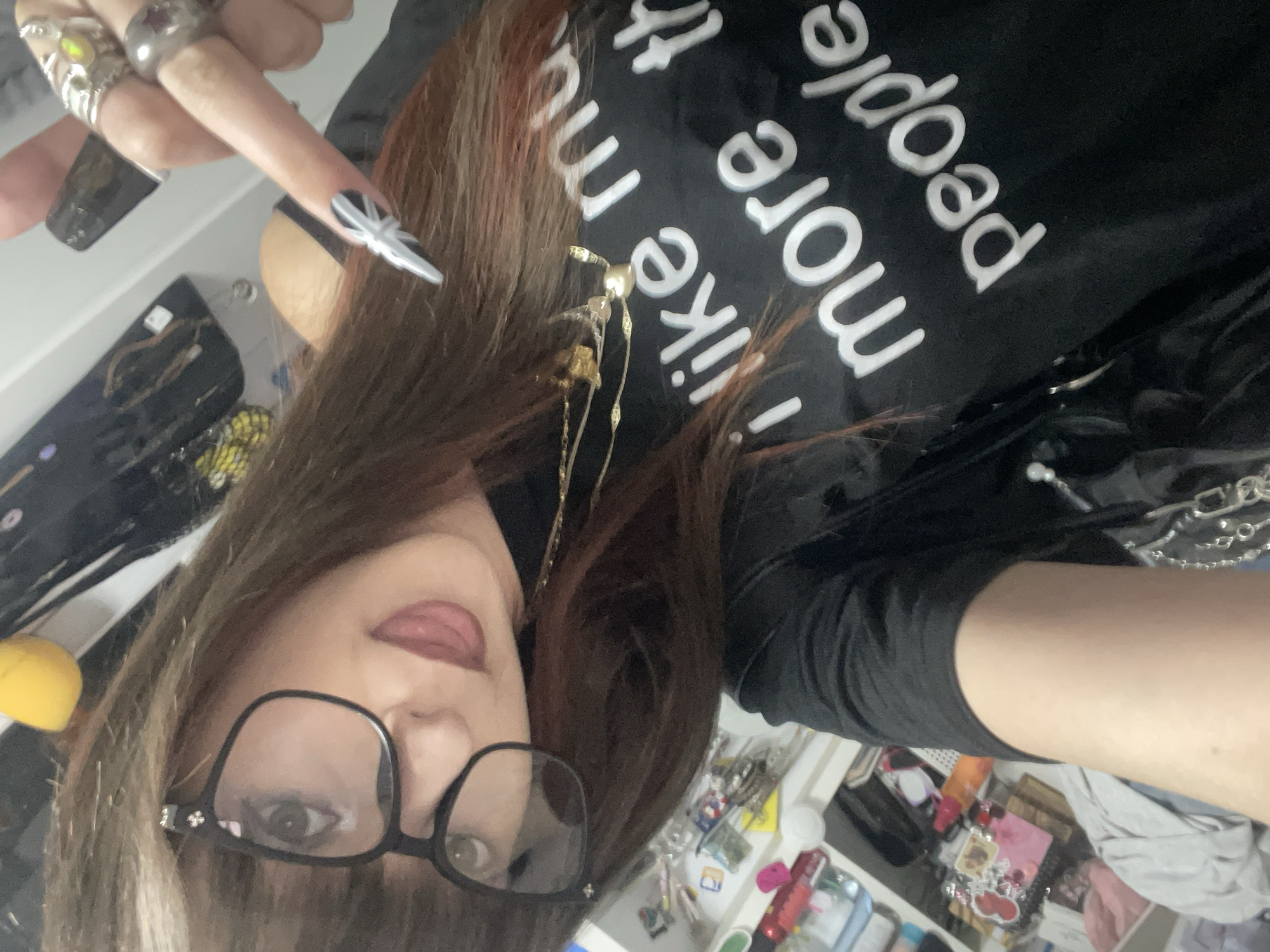 A woman wearing black-framed glasses and a black shirt with the words "more people like me" in white is taking a selfie indoors. She is sticking out her tongue and displaying an elongated fingernail with black and white designs. She has a gold necklace and several rings on her fingers. Her hair is brown with some red highlights. The background appears to be a cluttered room, possibly a bedroom or office. The photo does not reveal anything to indicate the city of Raanana, Israel.