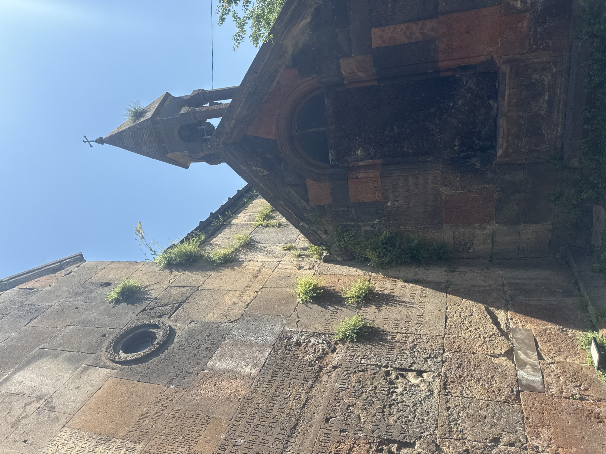 The image shows a section of the Bjni Fortress in Bjni, Armenia, featuring ancient stone structures bathed in sunlight under a clear blue sky. On the left, a large, weathered stone wall is covered in carved inscriptions. A circular window with decorative stonework is visible on this wall. To the right, a smaller stone building with a pointed bell tower stands, topped with a cross. The bell tower has an arched entrance and a dark, possibly wooden or metal door. Green vegetation grows in patches on the stone walls, suggesting age and weathering. The overall scene depicts historical architecture and an outdoor environment with strong natural lighting.
