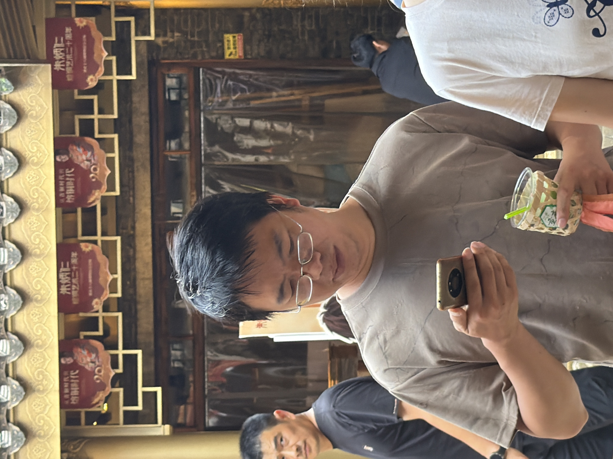 In Yuzhong District, China, a man in a t-shirt and glasses is looking at his phone, holding a cup with a straw in his other hand. He is in front of a store with signs featuring Chinese characters and images of people. Another man in a black t-shirt is visible to the left. In the background, a clear plastic curtain hangs over a doorway, revealing wooden structures. A brick wall is visible on the right, along with a small yellow sign. The scene appears to be outdoors or in a covered area with traditional Chinese architecture overhead.