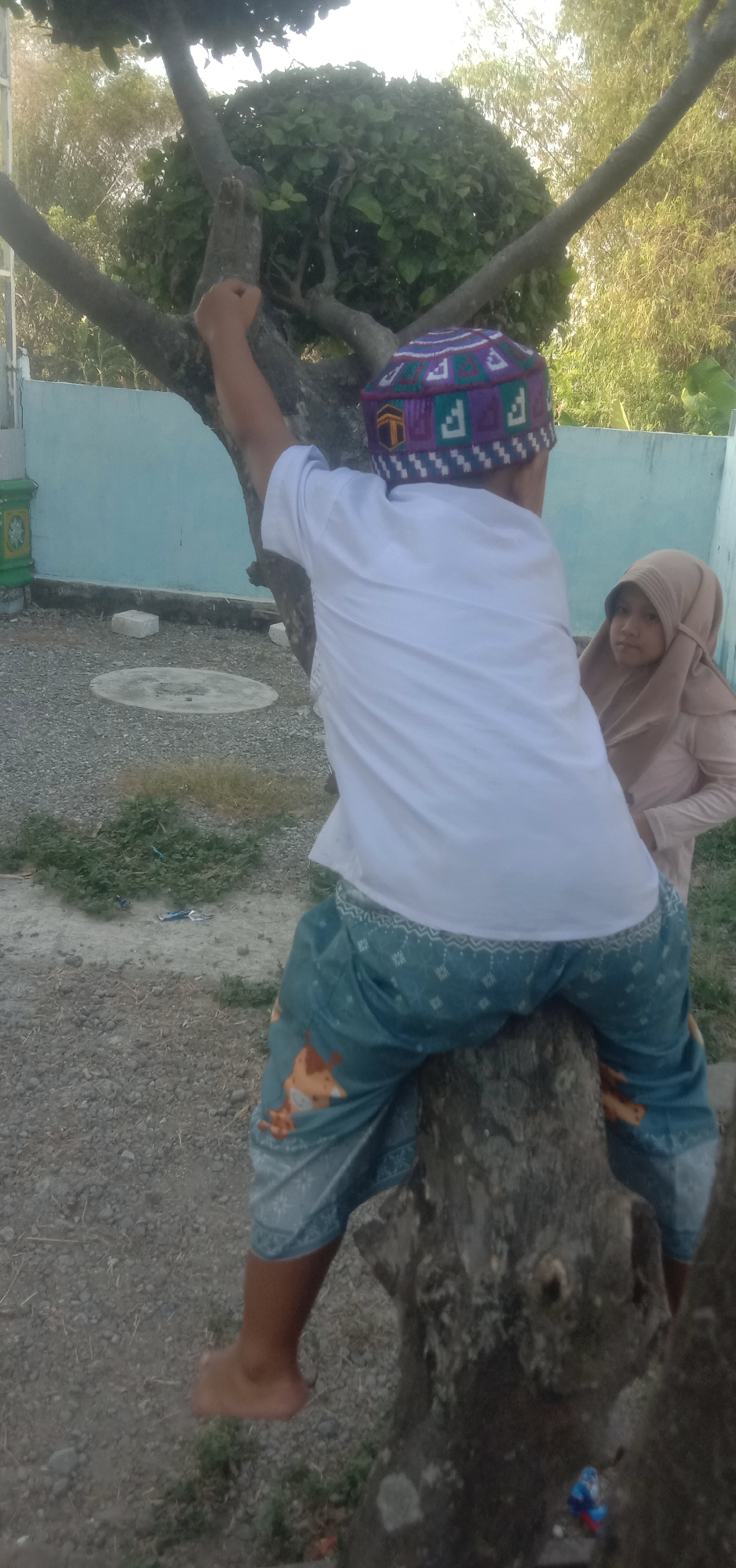 A boy wearing a white shirt and patterned pants sits on a tree trunk outdoors, facing away from the camera. He is wearing a purple and white patterned kufi. Beside him, a girl in a light brown hijab looks towards the camera. The environment appears to be a yard with gravel on the ground, scattered green grass, and a light blue wall in the background. Trees with green foliage are visible, and the lighting suggests it is daytime. The location is identified as Cangkringrandu, Indonesia.