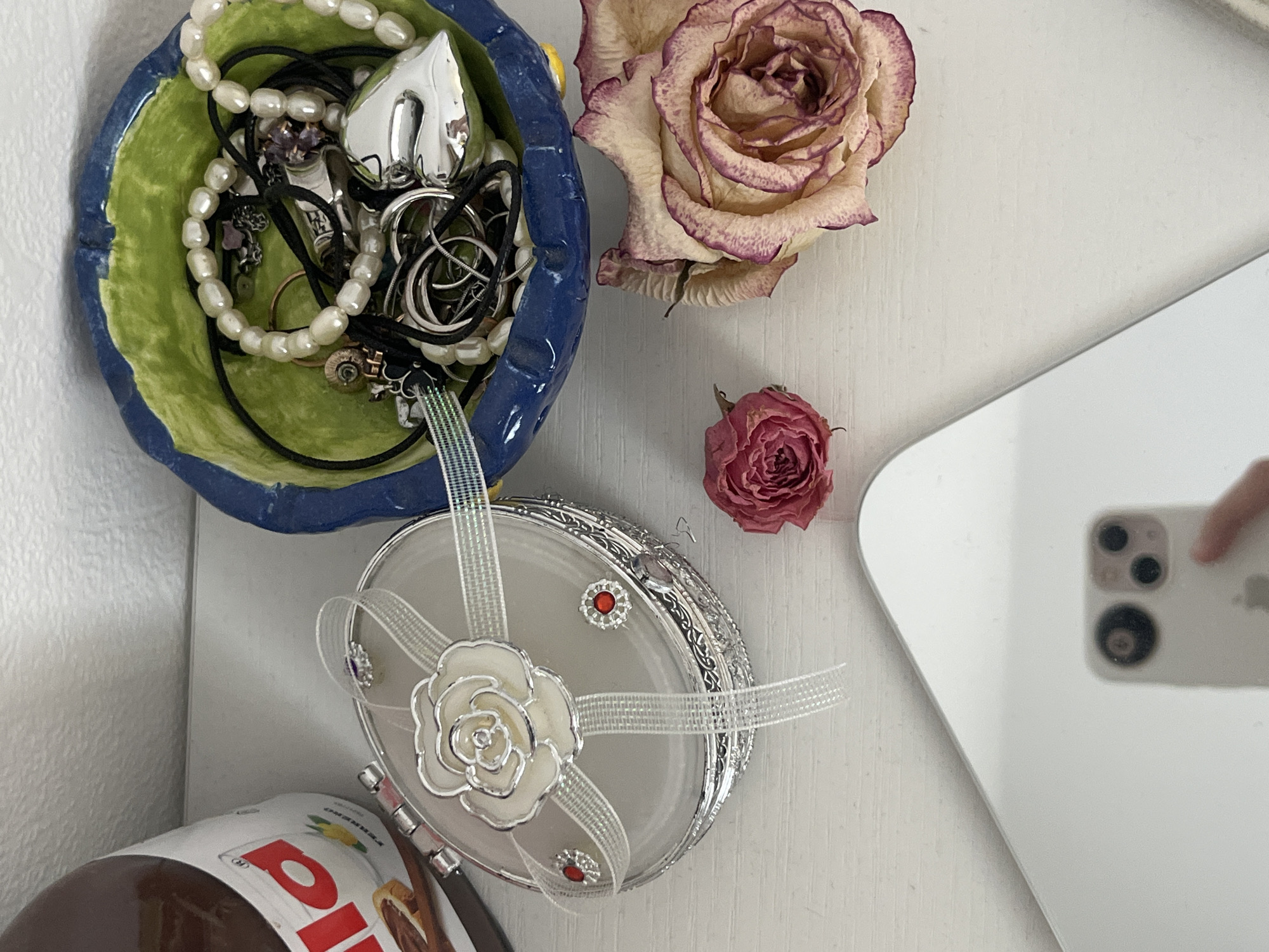 The image depicts a collection of decorative items arranged on a white surface. In the upper left corner, a partially visible jar of Nutella stands next to a silver jewelry box adorned with a white rose, ribbons, and red gems. To the right of the jewelry box, a blue and green ceramic bowl overflows with various pieces of jewelry, including necklaces, rings, and a heart-shaped pendant. Two dried roses, one small and pink, the other larger with faded pink and cream petals, lie near the jewelry bowl. In the foreground, a white smartphone is visible in the reflection of a large mirror, with a finger lightly touching the screen. The scene appears to be an indoor setting, possibly a vanity or dresser top, with a white wall in the background. There is no indication of a location in Moscow, Russia, or any activity that could be linked to that city.