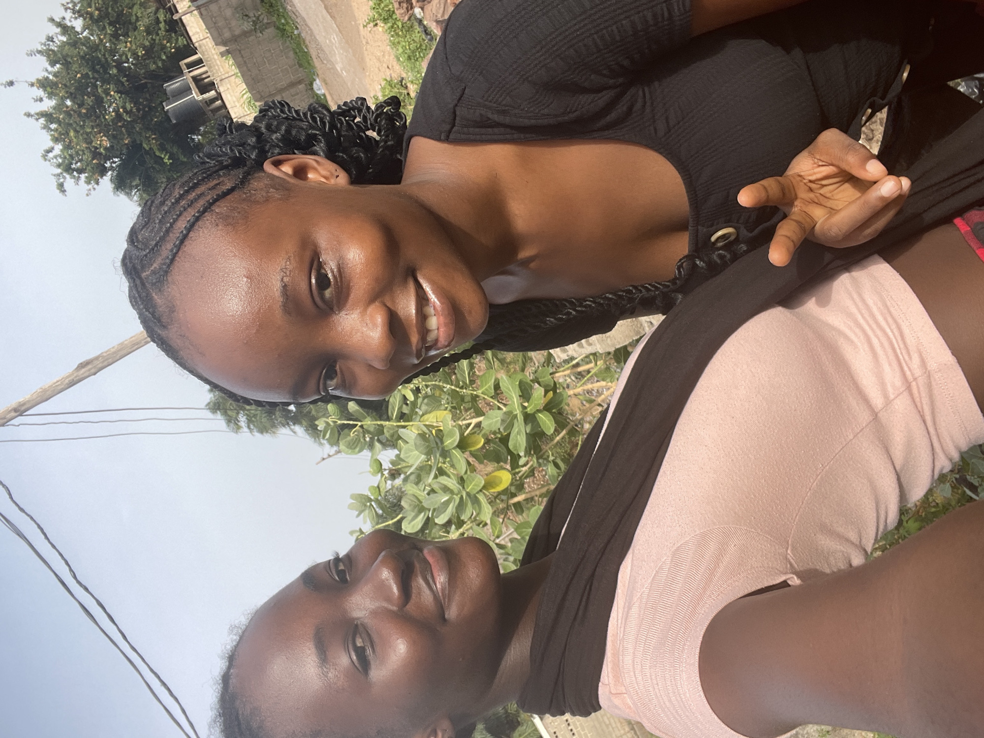 Two young women with dark skin are posing for a photograph in an outdoor setting, under a bright, likely overcast, daytime sky. The image appears to be a selfie.
The woman positioned closer to the camera and on the left side of the frame has braided hair styled with cornrows and twists. She is wearing a black ribbed top and is smiling broadly, looking directly at the camera. Her left hand is visible, making a gesture with her fingers.
The second woman, positioned slightly behind and to the right, has dark, braided hair and is wearing a light pink t-shirt with a black fabric draped around her neck or shoulders. She has a calm expression, looking slightly past the camera.
The background features lush green foliage, including trees and bushes, and a light-colored building constructed from concrete blocks. Thin utility wires are visible against the sky in the upper left. The ground appears to be dirt or earth. The bright natural lighting indicates it is daytime. No visible text is present. The scene's elements are consistent with an outdoor environment in Lokoja, Nigeria.