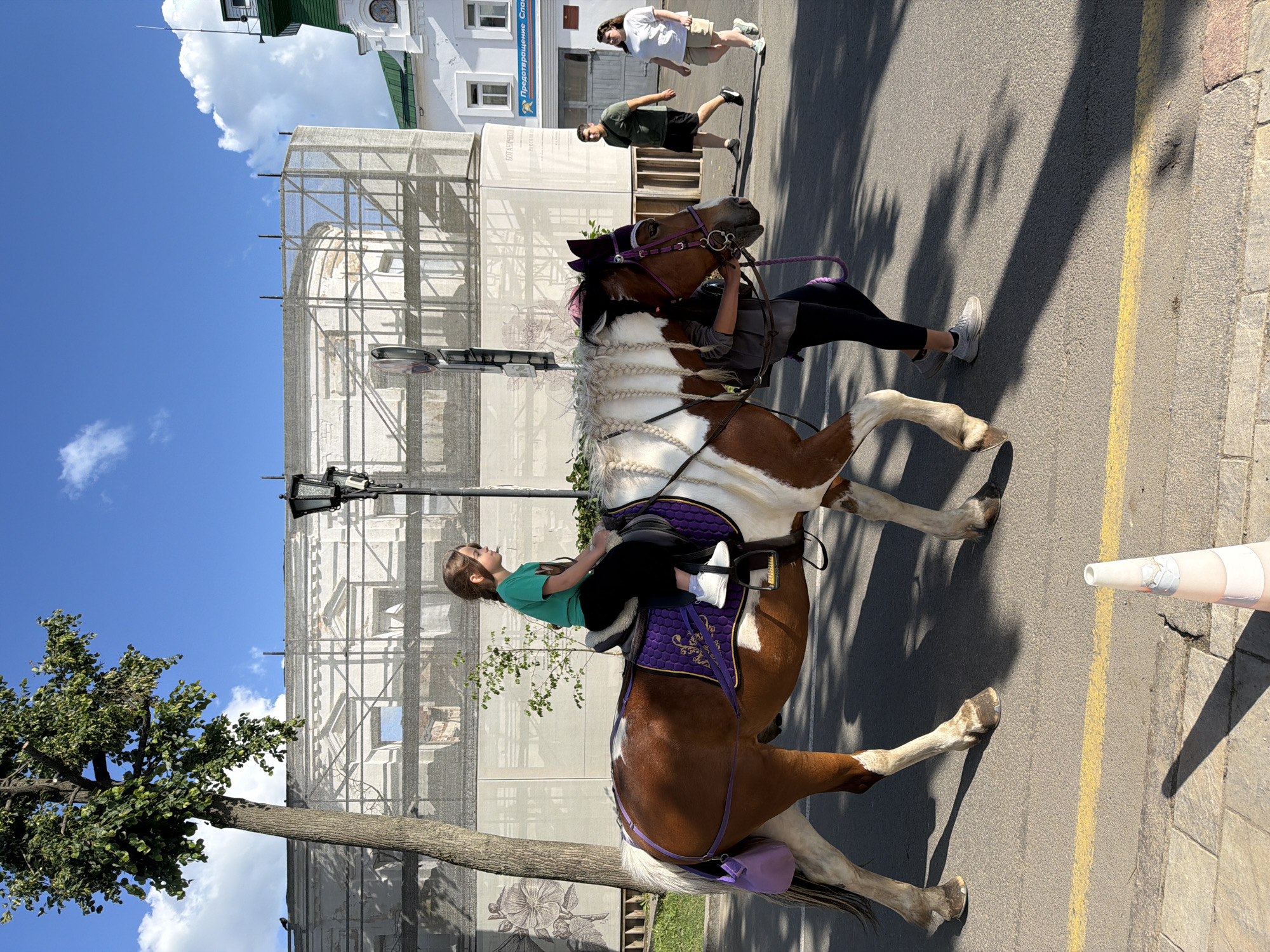A young girl with brown hair, wearing a green t-shirt and black pants, is riding a brown and white pinto pony. The pony has a purple saddle pad with gold trim and a purple bridle. The pony is being led by a person wearing a gray shirt and black pants, with dark hair. They are walking on an asphalt road with a yellow line, next to a sidewalk made of gray paving stones. In the background, a large building is covered in scaffolding and a white tarp, with a prominent street lamp. To the right, two adults are walking on the sidewalk. The taller adult, a woman with long dark hair, wears a white t-shirt and khaki shorts. The shorter adult, a man with short dark hair, wears a dark green t-shirt and black shorts. The scene is set on a sunny day with a blue sky and scattered white clouds, in the city of Suzdal, Russia.