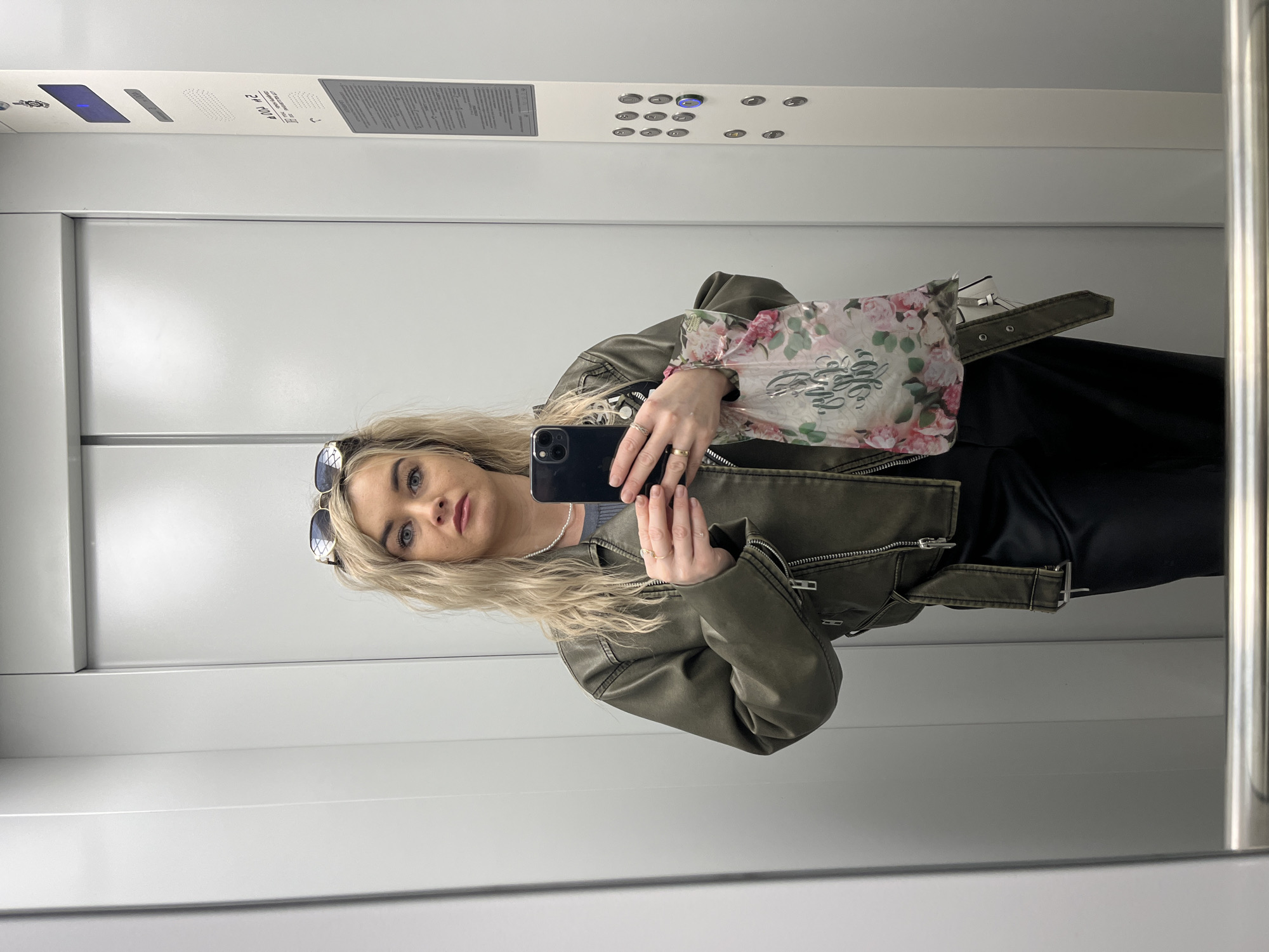 A woman is taking a selfie in an elevator. She is wearing a green leather jacket, a black skirt or pants, and sunglasses on her head. She is holding a black smartphone in her hands. The elevator interior is white with a metal control panel at the top. No information is available to confirm if this image was taken in the city of Chaikovsky, Russia.