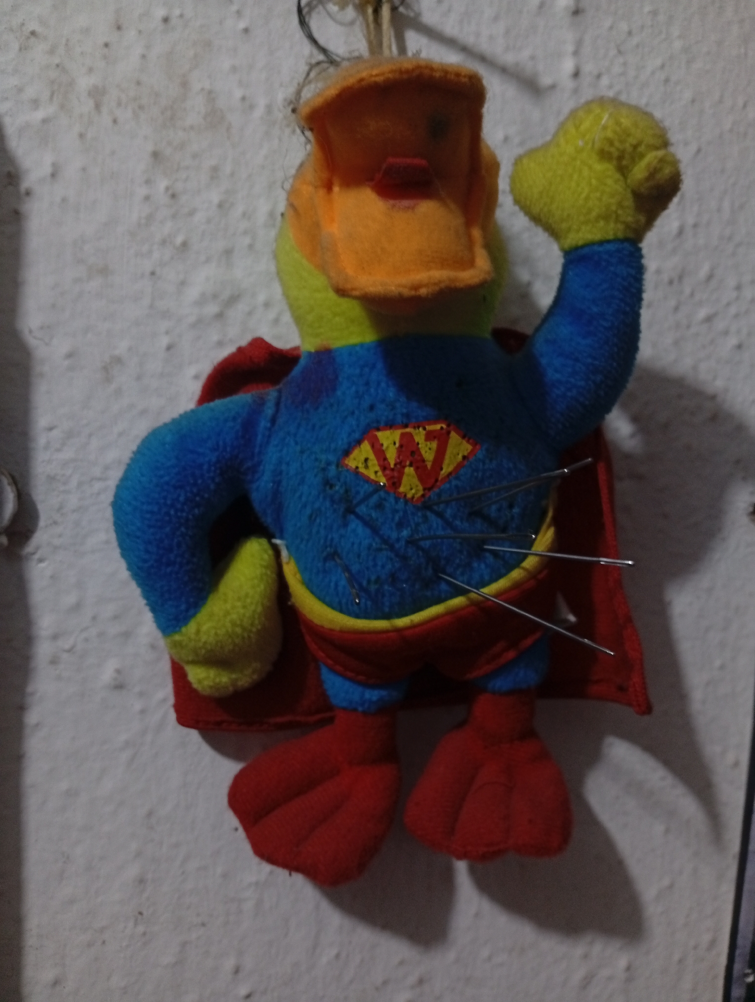 A plush toy depicting a duck dressed as a superhero is pinned to a white wall. The duck has a yellow head, an orange beak, a blue torso with a "W" emblem in a yellow diamond, a red cape, and red feet. Several sewing needles are stuck into the torso of the toy. The wall is textured and appears to be indoors. The location of the photograph is not discernible as Lokoja, Nigeria.