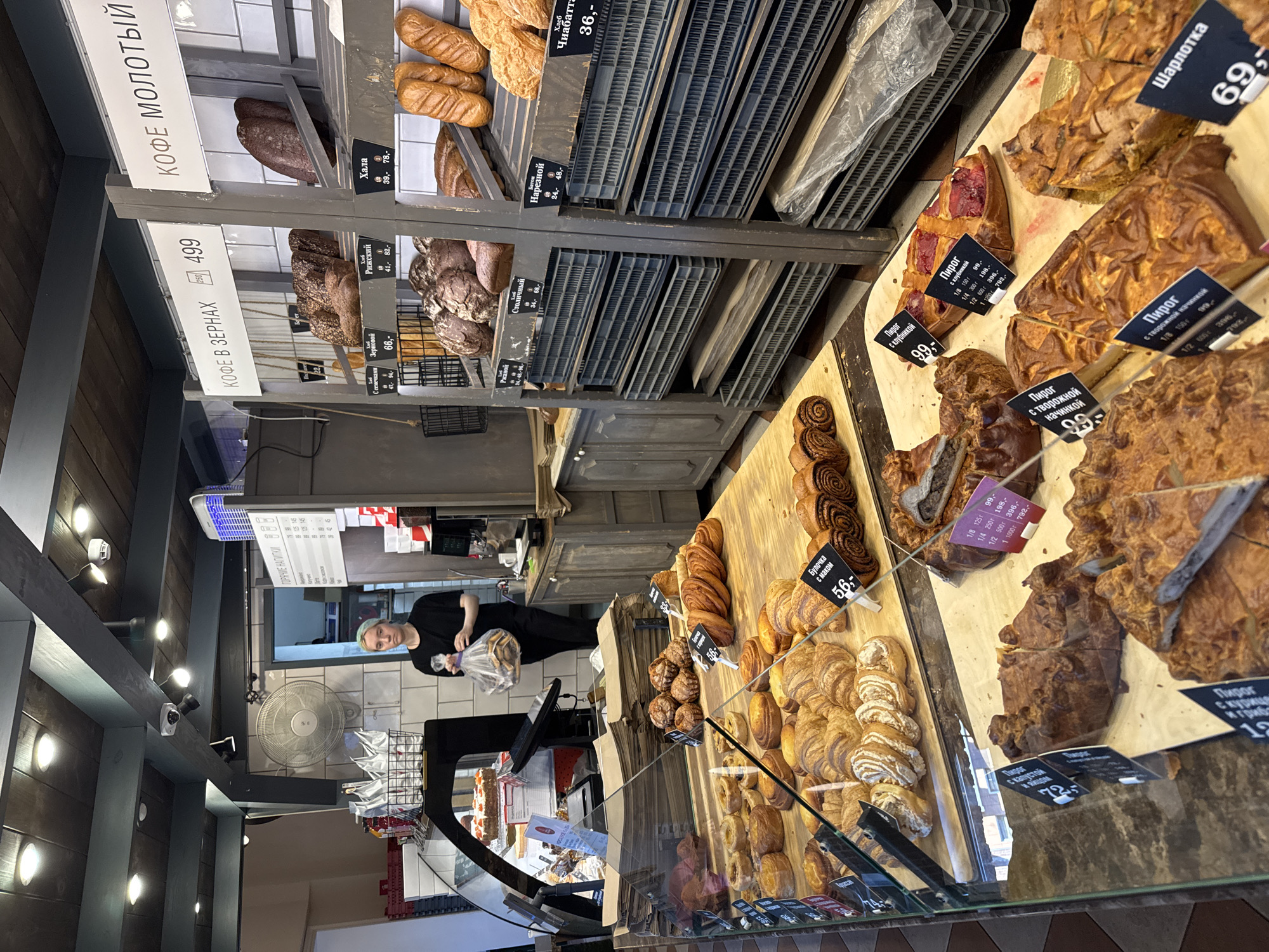 This image captures a view inside a bakery. Several shelves are laden with various baked goods, including loaves of bread, pastries, and pies. Some of the items are displayed in plastic crates. There are price tags visible with Russian text and prices. In the foreground, a display case shows more pastries.
A woman wearing a black shirt is visible in the background, holding a plastic bag filled with bread. She appears to be a customer or an employee. She is standing in front of a counter with a fan and some shelving.
The lighting suggests it could be daytime, possibly late afternoon, given the interior setting of a shop. The overall atmosphere seems to be one of a busy retail environment.
Visible text includes product names and prices in Russian, such as "КОФЕ В ЗЕРНАХ" (Coffee beans), "Хлеб Рижский" (Riga bread), "Шарлотка" (Charlotte cake), and "Пирог с клубникой" (Strawberry pie), among others.