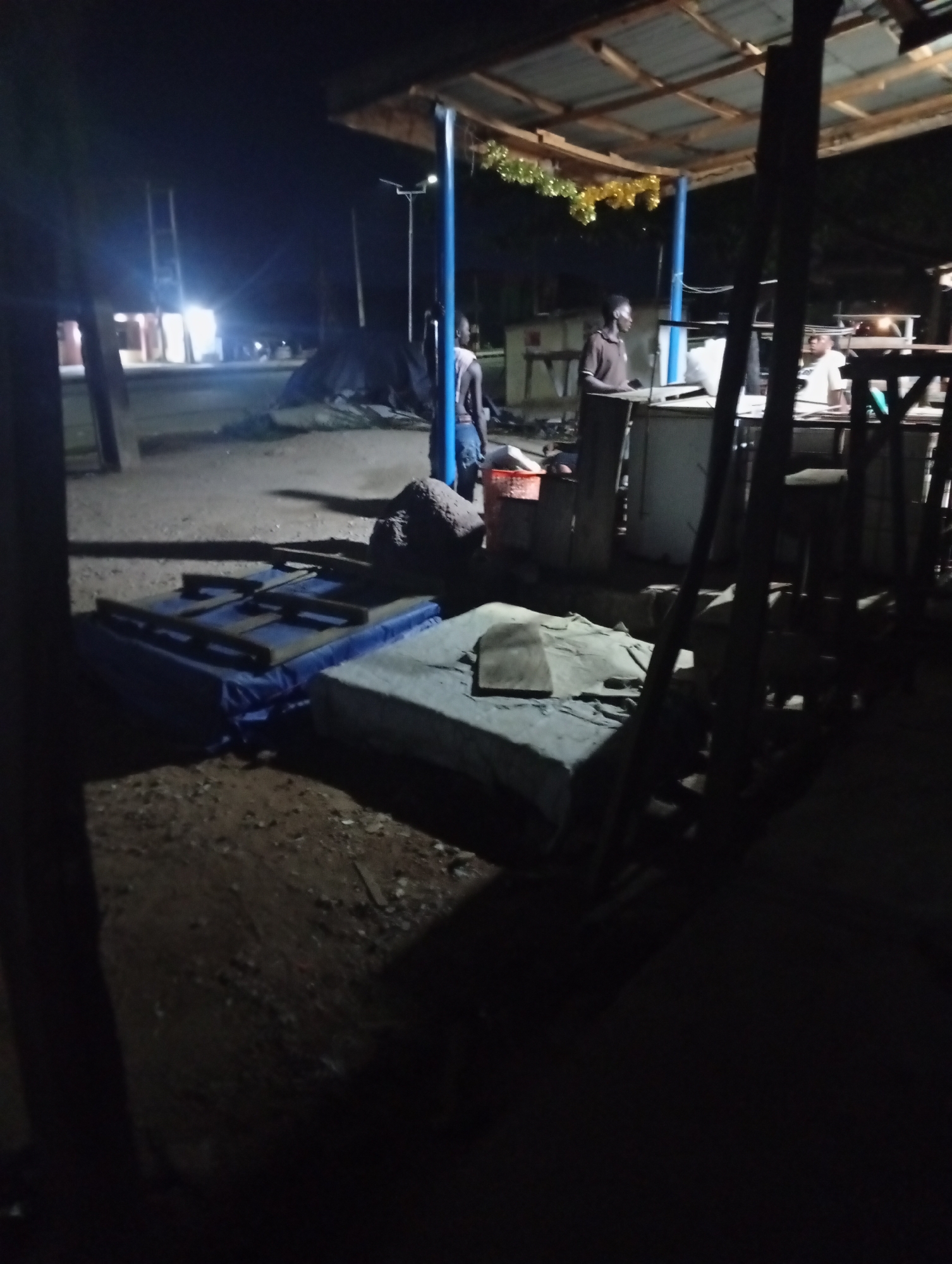 The image shows a nighttime scene in Adankolo, Nigeria. Under a wooden roof structure decorated with tinsel, three men stand near a makeshift bar or counter. Outside, two padded objects, possibly mattresses, lie on the ground. A tarpaulin-covered pile sits further back. Street lights illuminate the scene in the background. The environment is mostly dark with a dirt ground visible.