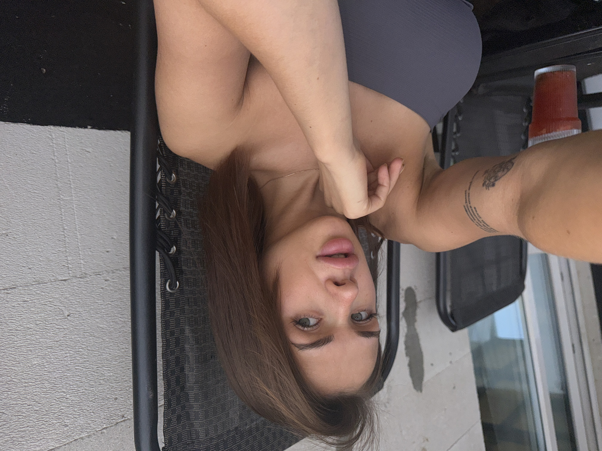 The image shows a young woman with brown hair and green eyes, sitting in a black outdoor chair. She is wearing a grey strapless top. Her left arm is extended towards the camera, and she has a tattoo on her upper arm. There is a red drink in a plastic cup to her left. The background consists of a white wall and some vertical blinds or windows. The scene appears to be outdoors, possibly on a balcony or patio, given the outdoor furniture and wall texture. The location is identified as Lobanovo, Russia.