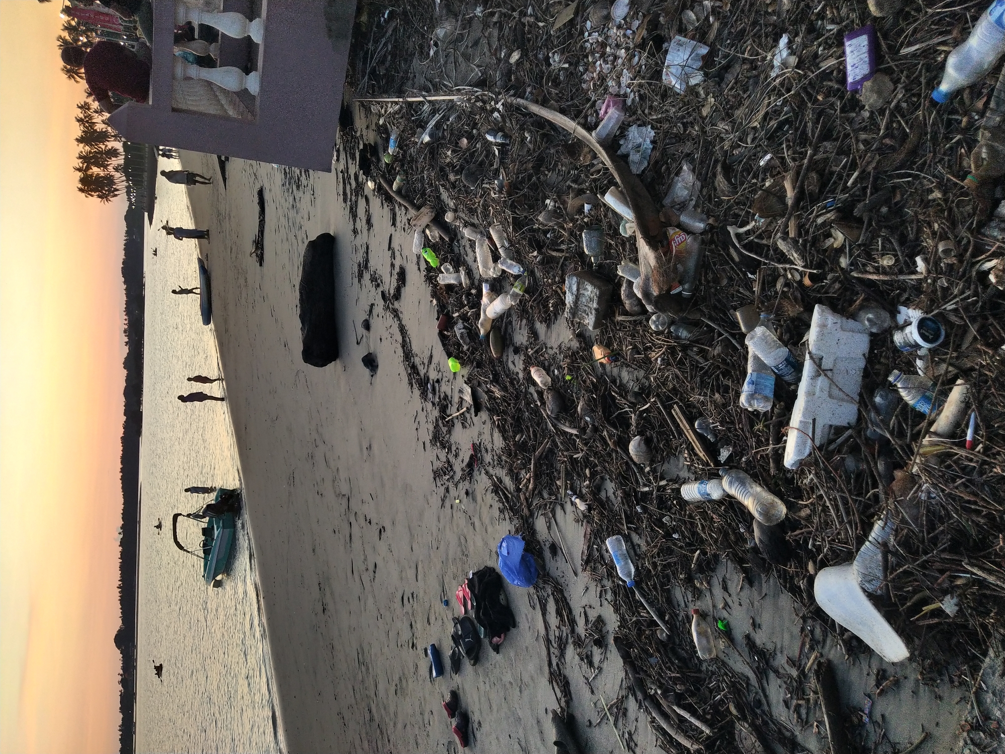The image depicts a polluted beach in Badem, India. Numerous plastic bottles, pieces of styrofoam, and other debris are scattered among natural materials like branches and leaves along the shoreline. A small boat is visible in the water near the beach. Several people are seen walking on the sandy shore. The scene is lit by the warm light of the setting sun, casting a golden hue over the sky and water.