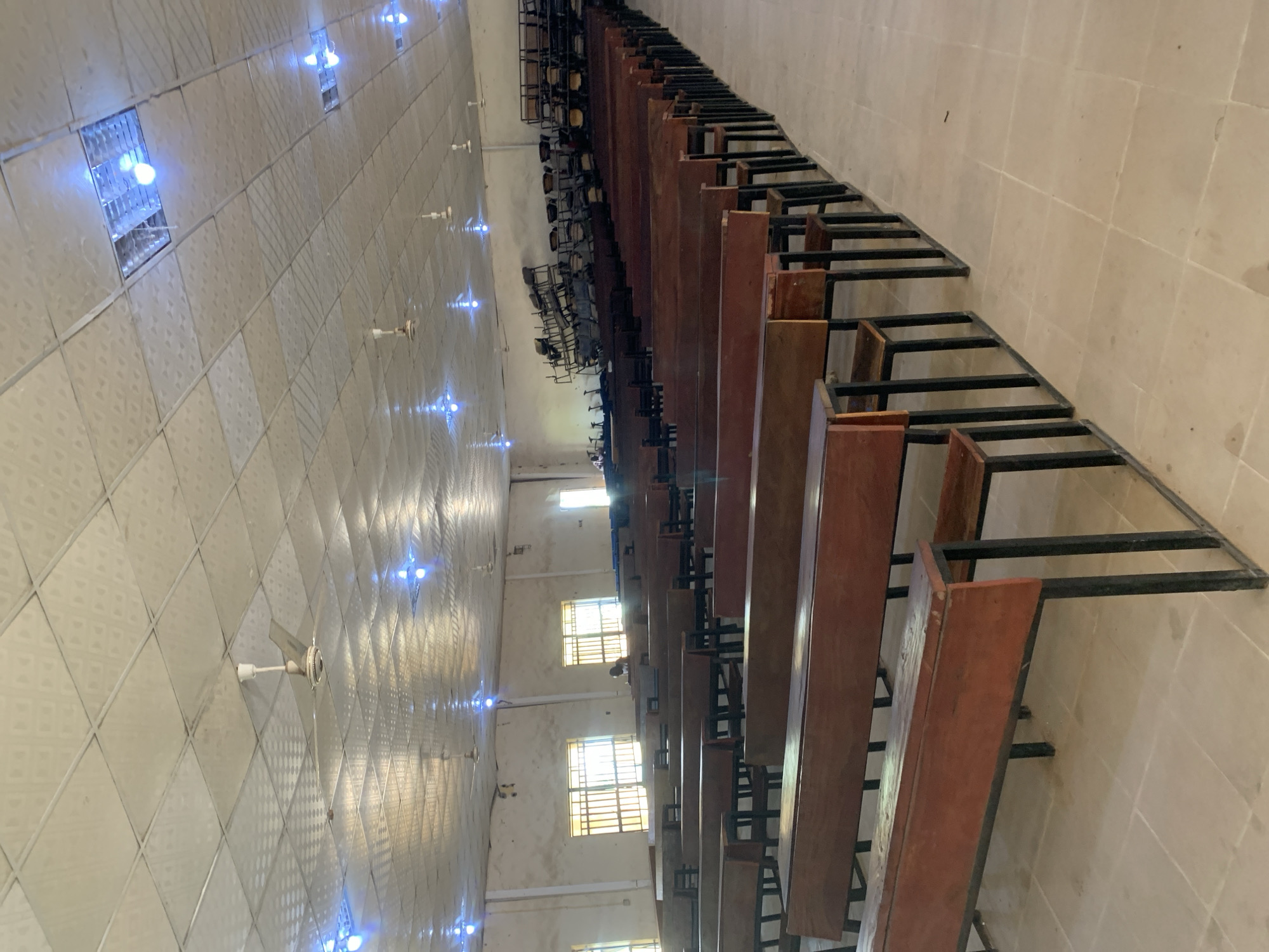 The image depicts the interior of a lecture hall or auditorium, likely located in Jalingo, Nigeria. The hall is furnished with rows of wooden benches, arranged in a semi-circular fashion, facing towards the front where chairs are stacked on elevated platforms. The ceiling is a drop ceiling with a textured pattern, punctuated by fluorescent lights and ceiling fans. Natural light enters through barred windows on the side walls. A single individual is visible in the distance, seated at one of the benches near the windows. The floor is tiled in a neutral color. The overall impression is one of a functional, though perhaps dated, educational space.