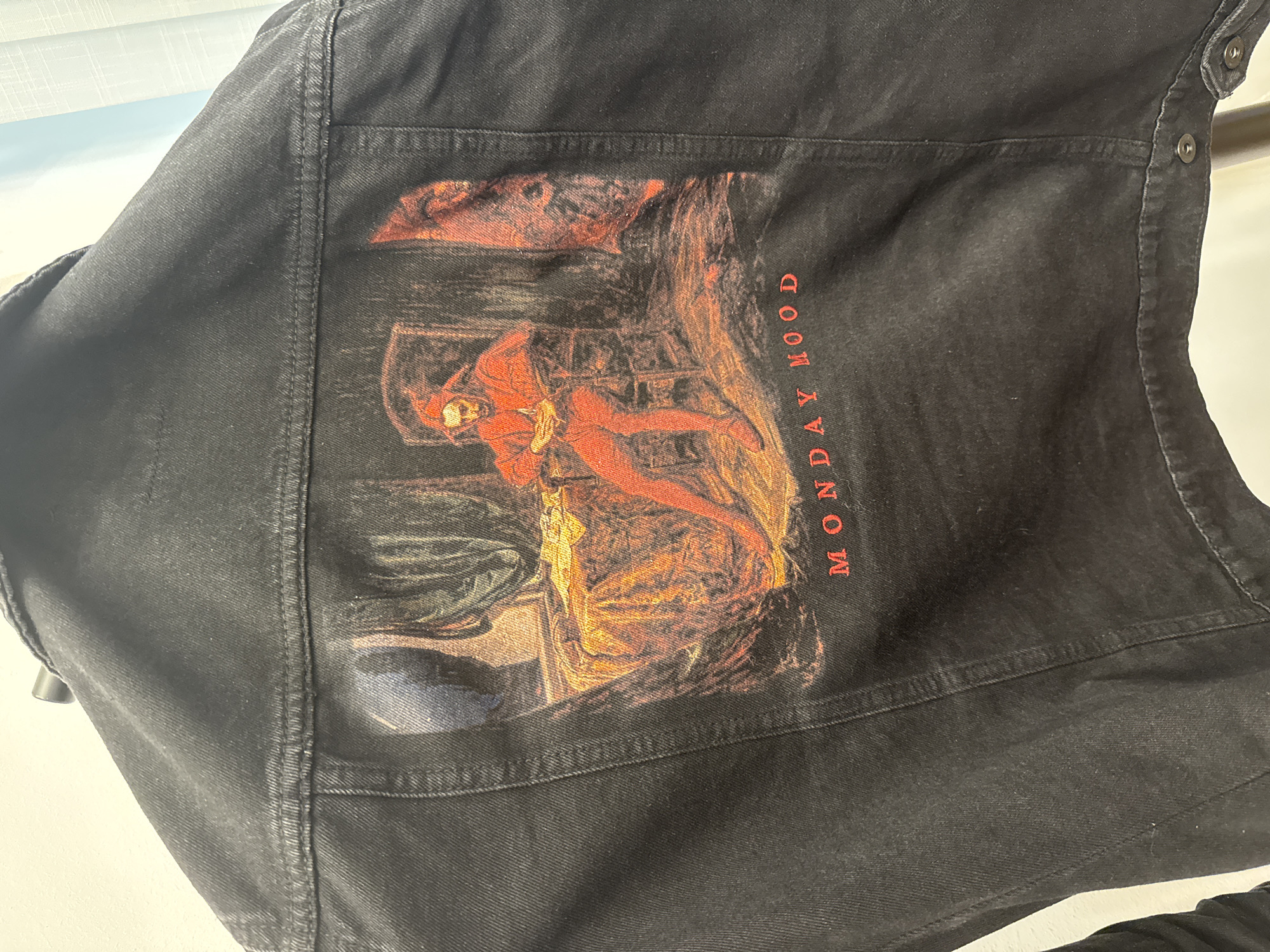 A black denim jacket, possibly a vest due to the visible side fasteners, is displayed at a slight angle. The jacket shows signs of wear, with subtle fading on its black fabric and prominent double stitching along its seams. Several metal snap buttons are visible on the right side.
The main focal point is a large, rectangular graphic printed on the back of the jacket. This artwork features a figure, seemingly female, rendered in a classical or pre-Raphaelite style. The figure is depicted in a reclining or lounging pose, dressed in flowing reddish-orange and golden-yellow drapery, which is brightly illuminated. The background of the artwork is darker, with muted tones of blue, green, and brown, suggesting an intimate, indoor setting such as a chamber or bedroom. The figure's posture implies a mood of contemplation or relaxation.
To the right side of the printed artwork, oriented vertically, is the text "MONDAY MOOD" in distinct red capital letters.
The photograph is well-lit, providing clear detail of the jacket's texture and the printed graphic. The background behind the jacket is blurry and light-toned, indicating an indoor setting.
