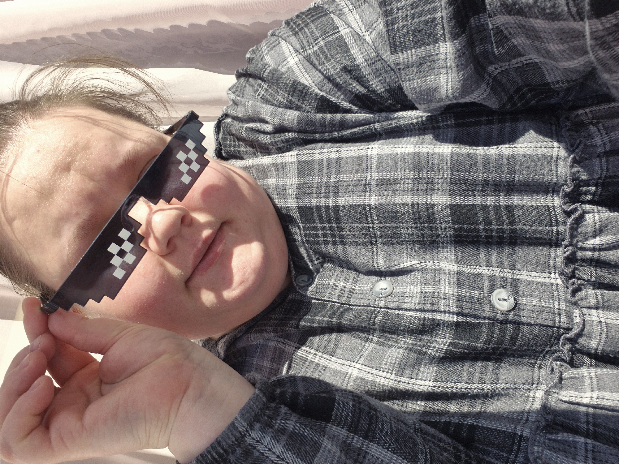 A person with fair skin is lying down, wearing pixelated sunglasses and a grey and black plaid shirt. The person's left hand is holding the sunglasses. The background is a light, neutral color, possibly a blanket or sheet. The location is unknown and cannot be determined to be Chaikovsky, Russia based on the visual information provided.