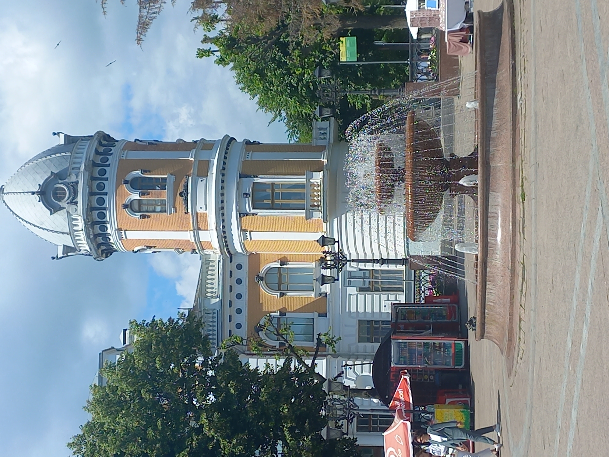 This image shows a fountain in a public square in front of a historic building with a distinctive tower. The building has yellow brickwork and white architectural details. The tower is capped with a dome and features arched windows. In the foreground, a multi-tiered fountain sprays water. To the left of the fountain, there are two street lamps and a kiosk selling drinks. Two people are walking in the square. The sky is blue with some clouds, and trees are visible in the background. This scene is located at the Museum of Civil Aviation History in Ulyanovsk, Russia.
