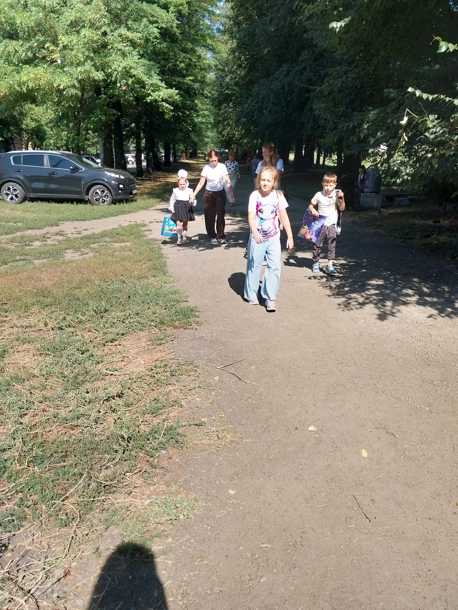 In Dnipro, Ukraine, a group of people, including children, walk along a dirt path bordered by lush green trees and dry grass on a sunny day. A dark gray SUV is parked to the left of the path. The children appear to be walking from right to left, carrying colorful bags. The focus of the image is on the children and their interactions as they move through the park-like setting.