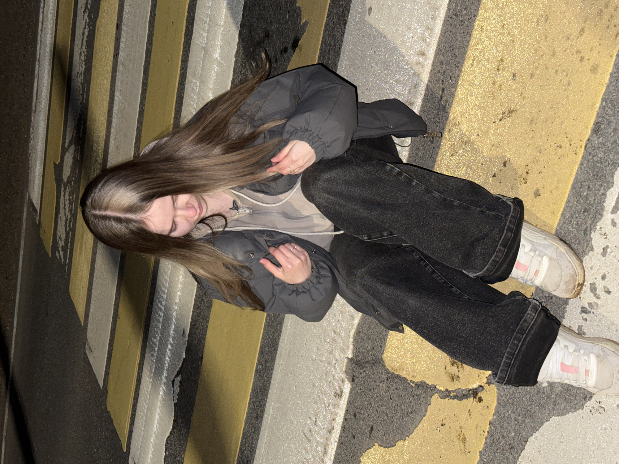 A young woman is sitting on a pedestrian crosswalk. She has long brown hair and is wearing a gray puffer jacket, a light-colored hooded sweatshirt, dark jeans, and white sneakers. The crosswalk has yellow and gray stripes. The photo was taken from an overhead angle. The exact location in the city of Unknown, Russia is difficult to determine without more information, but the presence of a pedestrian crosswalk indicates an urban area.