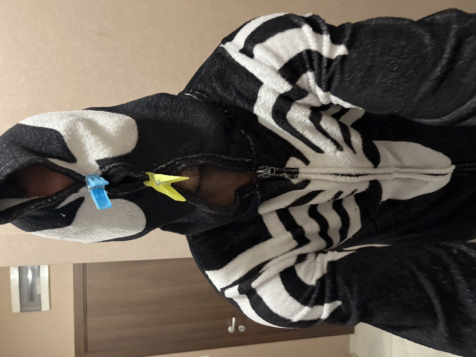 A person wearing a black and white fuzzy costume, resembling Venom from Spider-Man, is standing in a room. The costume has a hood with white eyes and black and white stripe detailing on the chest and arms. Two clothespins, one blue and one yellow, are clipped to the front of the costume, near the person's mouth area. The background consists of a beige wall and a closed wooden door with a silver handle on the left side. A white electrical panel is visible on the wall above and to the left of the door. The lighting appears to be artificial, casting soft shadows. No specific location within Sofia, Bulgaria, is identifiable from the image.