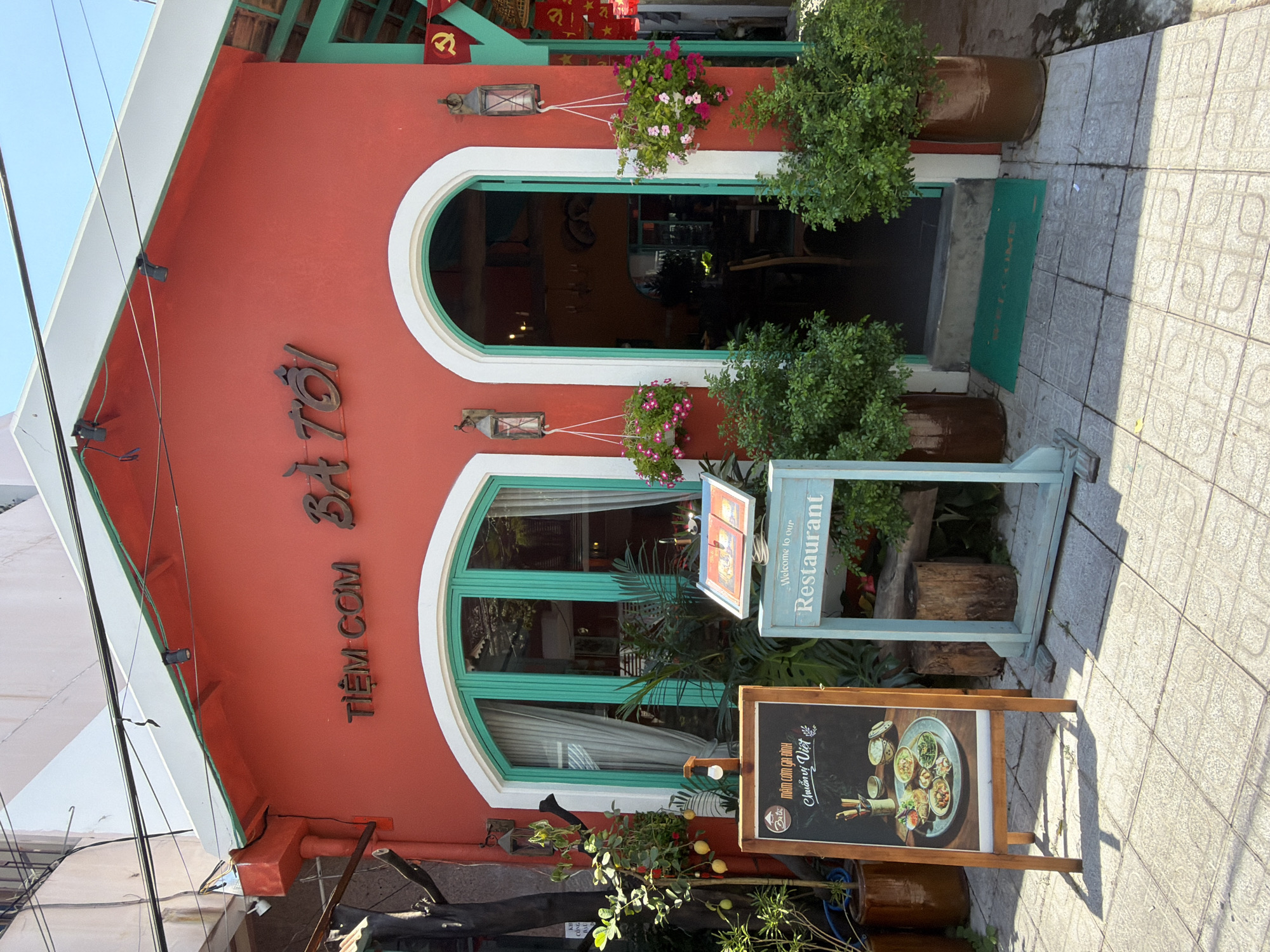 The image depicts a vibrant, red-fronted restaurant in Nha Trang, Vietnam, identified by the text "TIỆM CƠM BÀ TÔI" which translates to "Ba Toi's Rice Shop." The restaurant features teal-colored window frames and an arched doorway. A wooden easel displays a menu with an image of a Vietnamese dish. Next to it, a light blue wooden sign reads "Welcome to our Restaurant." Green plants in large pots flank the entrance, and hanging baskets filled with pink flowers add a decorative touch. Red banners with a yellow star, characteristic of the Vietnamese flag, are visible on the right side of the building. The ground is paved with light-colored square tiles, and sunlight casts shadows on the pavement. The overall scene is bright and inviting, suggesting a typical street view of a local eatery in the city.
