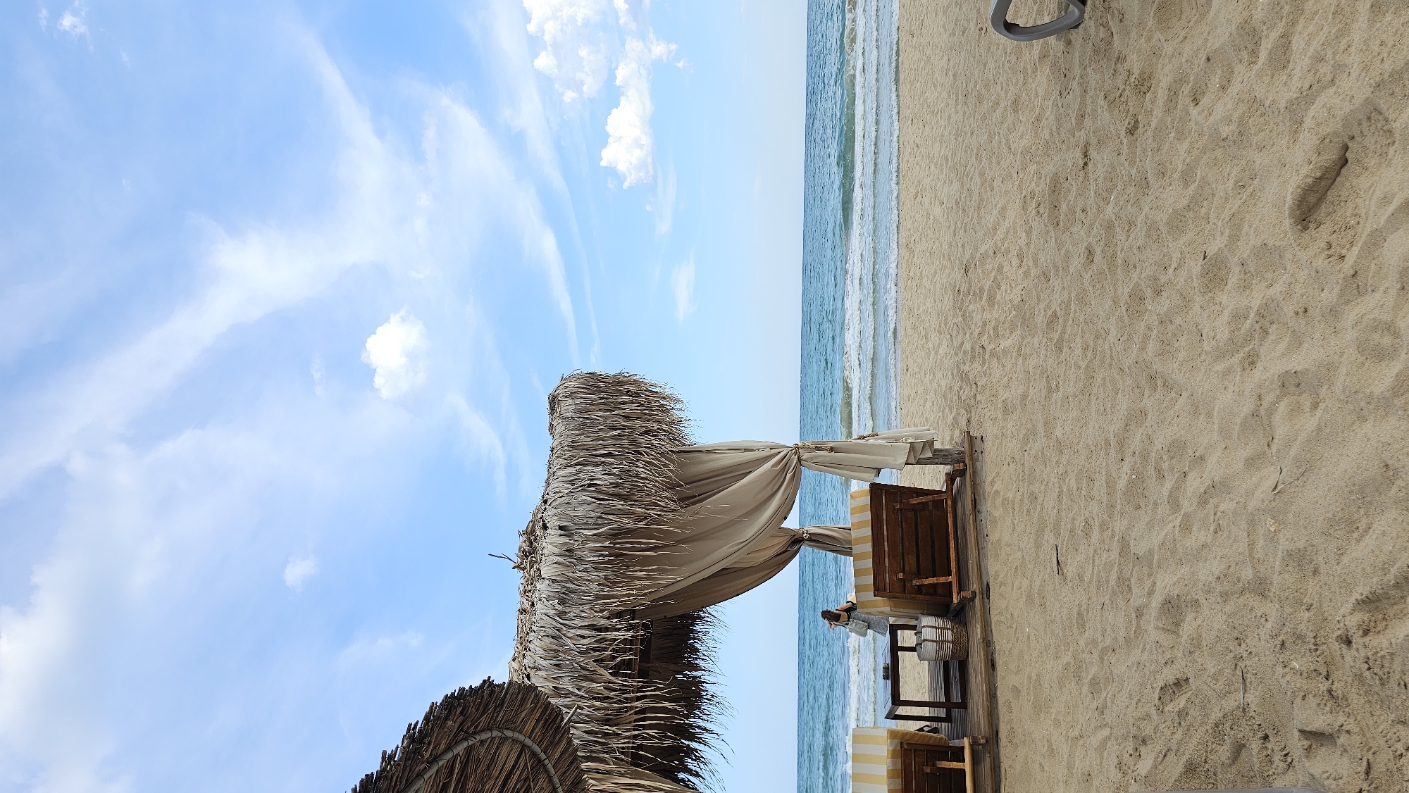 The image depicts a tranquil scene at a beachside establishment in Varna, Bulgaria. In the foreground, golden sand is marked with footprints, suggesting recent activity. To the left, a thatched-roof cabana provides shade, furnished with two striped lounge chairs and a small wooden table. Light-colored drapes hang from the cabana, adding to the relaxed atmosphere. A woman, seen from behind, stands near the table, holding a bag. Beyond the cabana, the calm, blue sea meets the horizon under a sky filled with wispy white clouds. The gentle waves create a soothing soundscape, contributing to the overall serene ambiance.