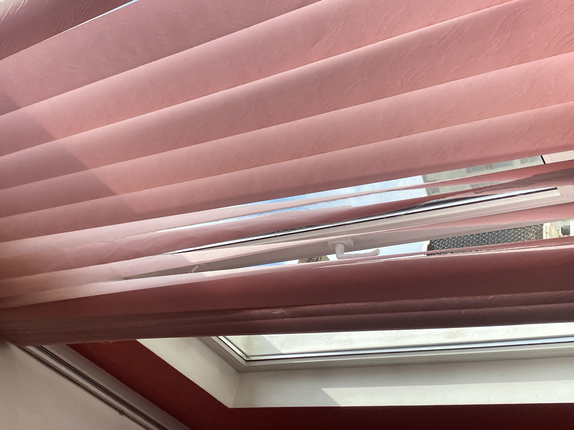 The image provides an upward-looking interior view through a partially opened skylight or roof window. The primary subjects are horizontal fabric blinds, which are light pink or rose in color and feature a subtle, textured, wavy pattern. Bright daylight illuminates the blinds, casting soft shadows on their lower surfaces.
Through the gaps in the partially open blinds, sections of a clear blue sky with some white clouds are visible, indicating sunny weather. A white window frame, likely made of uPVC, is prominent, and a white handle is visible on the right side, suggesting the window can be opened. Beyond the window and through the blinds, a glimpse of an exterior structure with a metallic or patterned surface can be seen, possibly a roof or ventilation unit of an adjacent building. Below the window, a section of a red-painted interior wall is visible, alongside white trim or a window sill.
No people or discernible activities are present in the static scene. The time of day is clearly daytime, characterized by bright sunlight. There is no visible text. The setting appears to be an interior space, likely in a residence or building in Troyan, Bulgaria.
