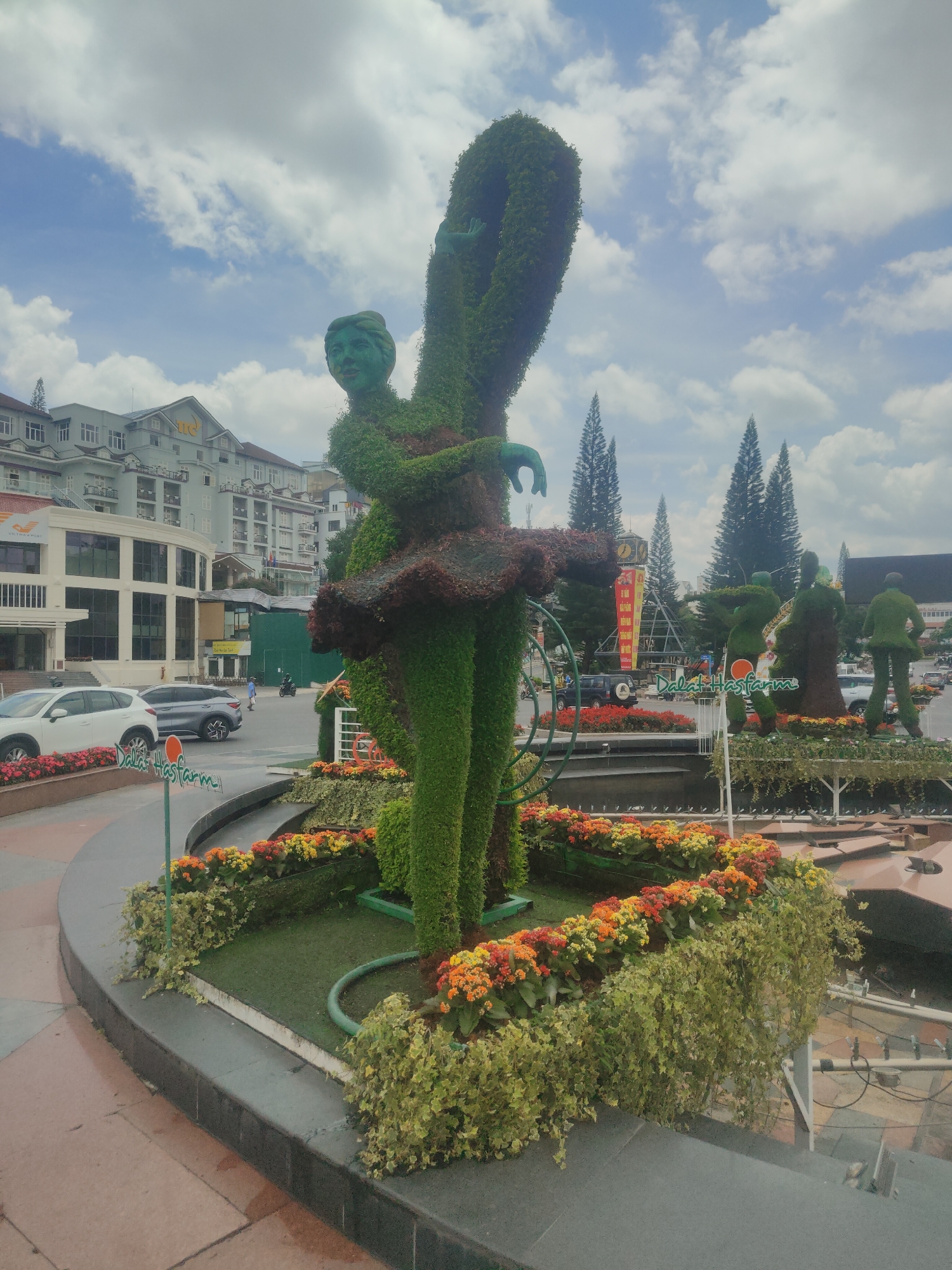 A tall, green topiary sculpture of a woman in a ballet pose stands in the center of a city square in Da Lat, Vietnam. The sculpture is adorned with a dark brown tutu and a green face. Surrounding the base of the sculpture are vibrant flowerbeds in shades of orange, red, and yellow, interspersed with lush green foliage. In the background, modern buildings with balconies and windows line the street, with several cars and a motorbike visible. Pine trees rise above the buildings under a cloudy sky. Another topiary sculpture depicting people is visible further in the background, along with a clock tower. A sign with "Dalat Hasfarm" is displayed in front of the flowerbeds.