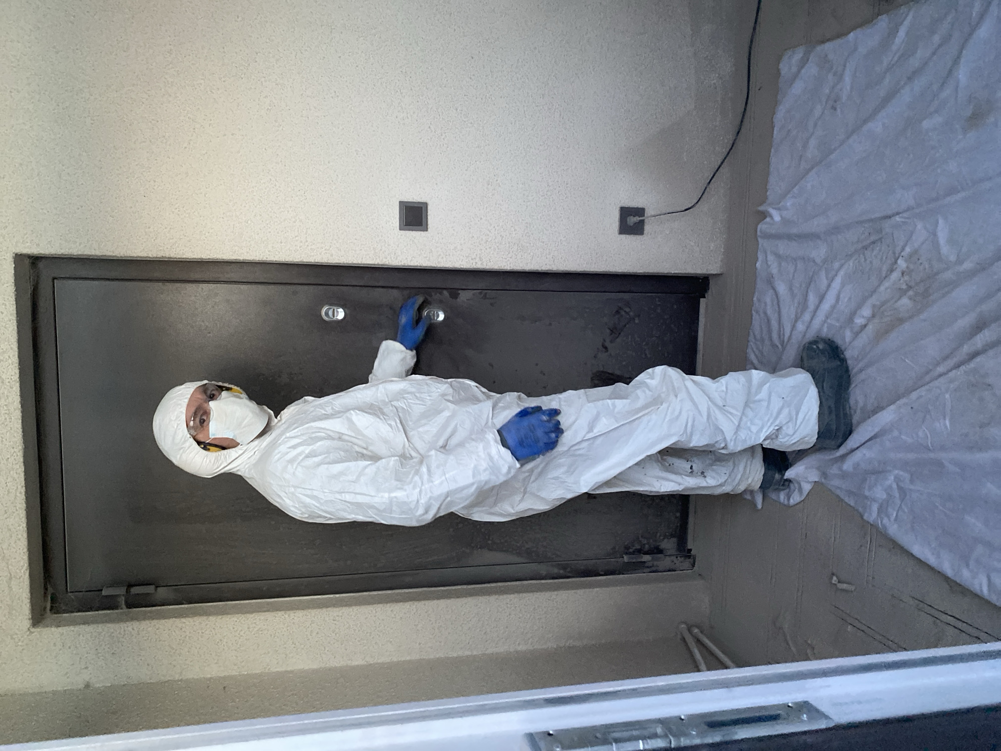 An individual is depicted in an indoor environment, identified as a location within the city of Chaikovsky, Russia. The person, viewed from a slight right-side angle, is standing near a dark metal door. They are fully clad in white personal protective equipment, consisting of a hooded coverall, clear safety glasses or goggles, a white respirator mask, yellow ear protection muffs, and blue protective gloves. Dark protective boots are visible on their feet. Their right hand is positioned near or on a silver-colored circular door handle/lock mechanism of the dark door. The door itself features visible hinges on its left side.
To the right of the door, the wall is light-colored and textured. Two square-shaped electrical components are affixed to this wall: an upper, dark-colored switch and a lower, dark-colored electrical outlet. A black power cord is plugged into the lower outlet, extending downwards and to the right, traversing over a light-colored, possibly white or light grey, protective sheet or tarp laid on the dusty concrete floor. The individual is standing on this protective sheet. On the far left of the image, a portion of a white door frame or an open white door is visible, revealing light-colored pipes against a wall in the background.