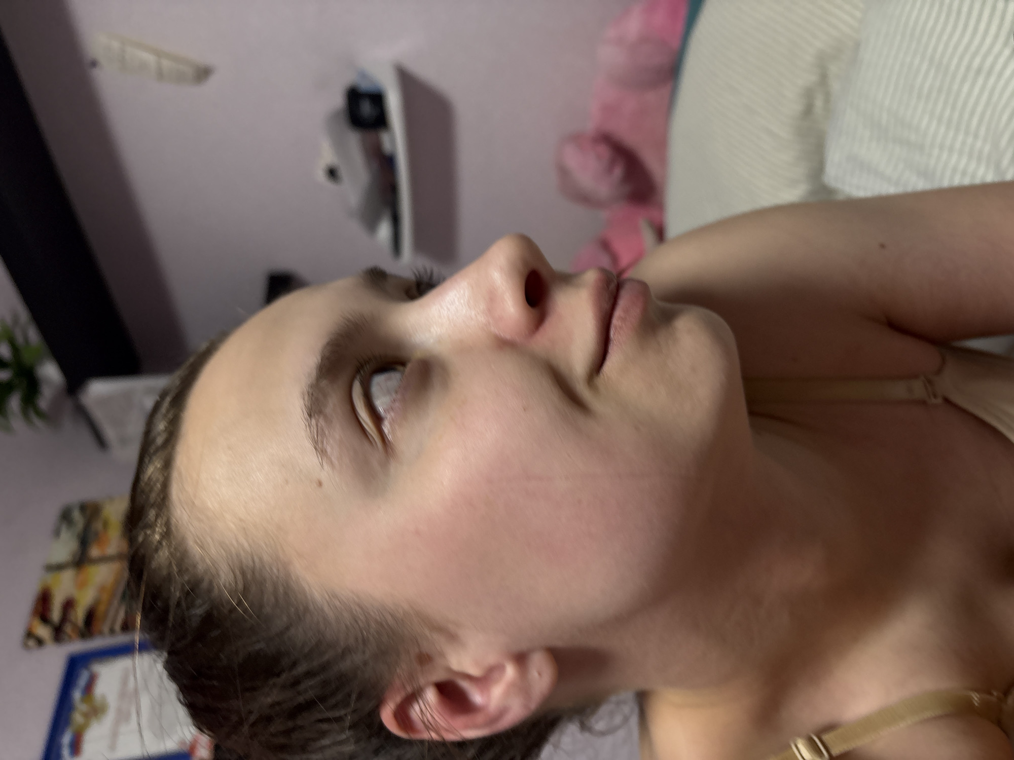 A side-view close-up shot of a fair-skinned woman with brown hair is featured. She is positioned on her back, with her head tilted upward. Her eyes are open, and her lips are gently closed. She has a visible bra strap on her shoulder. The background includes a bed with striped bedding, a pink teddy bear, a shelf with items, and a wall with decorations. The specific location of Прикамский, Russia, is not discernible from the image.