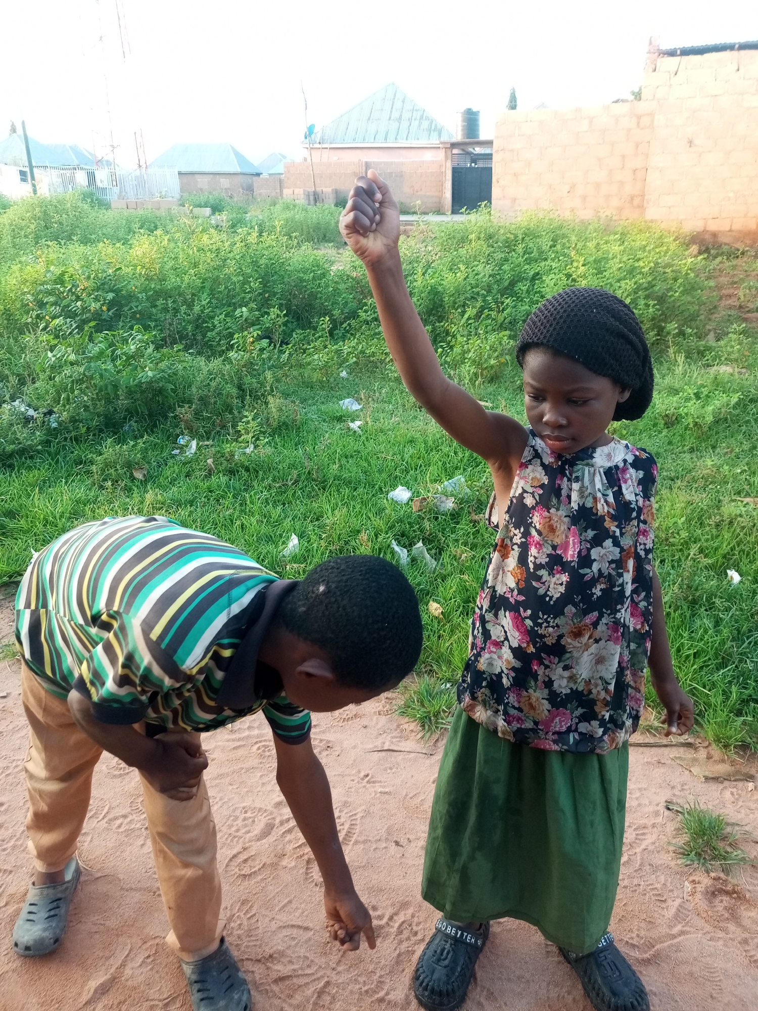 Two children, a boy and a girl, are in the foreground of the image, standing on a sandy patch of ground with grass and buildings in the background. The boy, on the left, is wearing a striped shirt and light-colored pants, and is bent over, appearing to examine something on the ground. The girl, on the right, is wearing a floral-patterned dress and a dark head covering, and has her right arm raised in the air. The environment appears to be an outdoor setting in Tempure, Nigeria, with the characteristic brick buildings and sparse vegetation visible. The lighting suggests it is daytime, possibly late afternoon due to the shadows.
