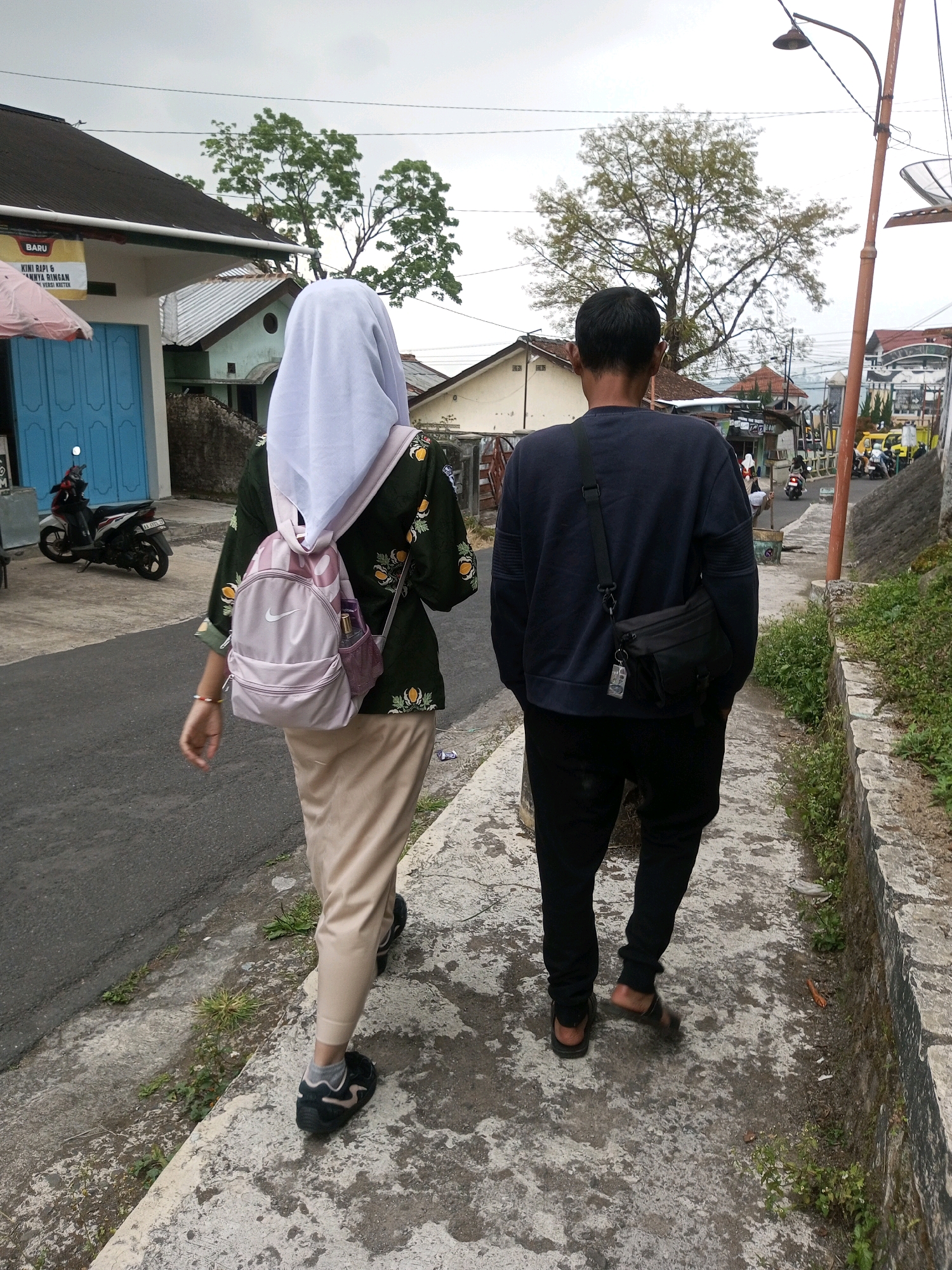 Two individuals, a woman and a man, walk away from the camera on a paved sidewalk. The woman, wearing a light-colored hijab, a dark green patterned long-sleeved shirt, and beige pants, carries a light pink backpack. The man, wearing a dark blue long-sleeved shirt and black pants, has a black crossbody bag. They are walking on a sidewalk that runs alongside a road. The environment appears to be a residential area in Krasak, Indonesia, with houses and trees visible in the background. The sky is overcast.