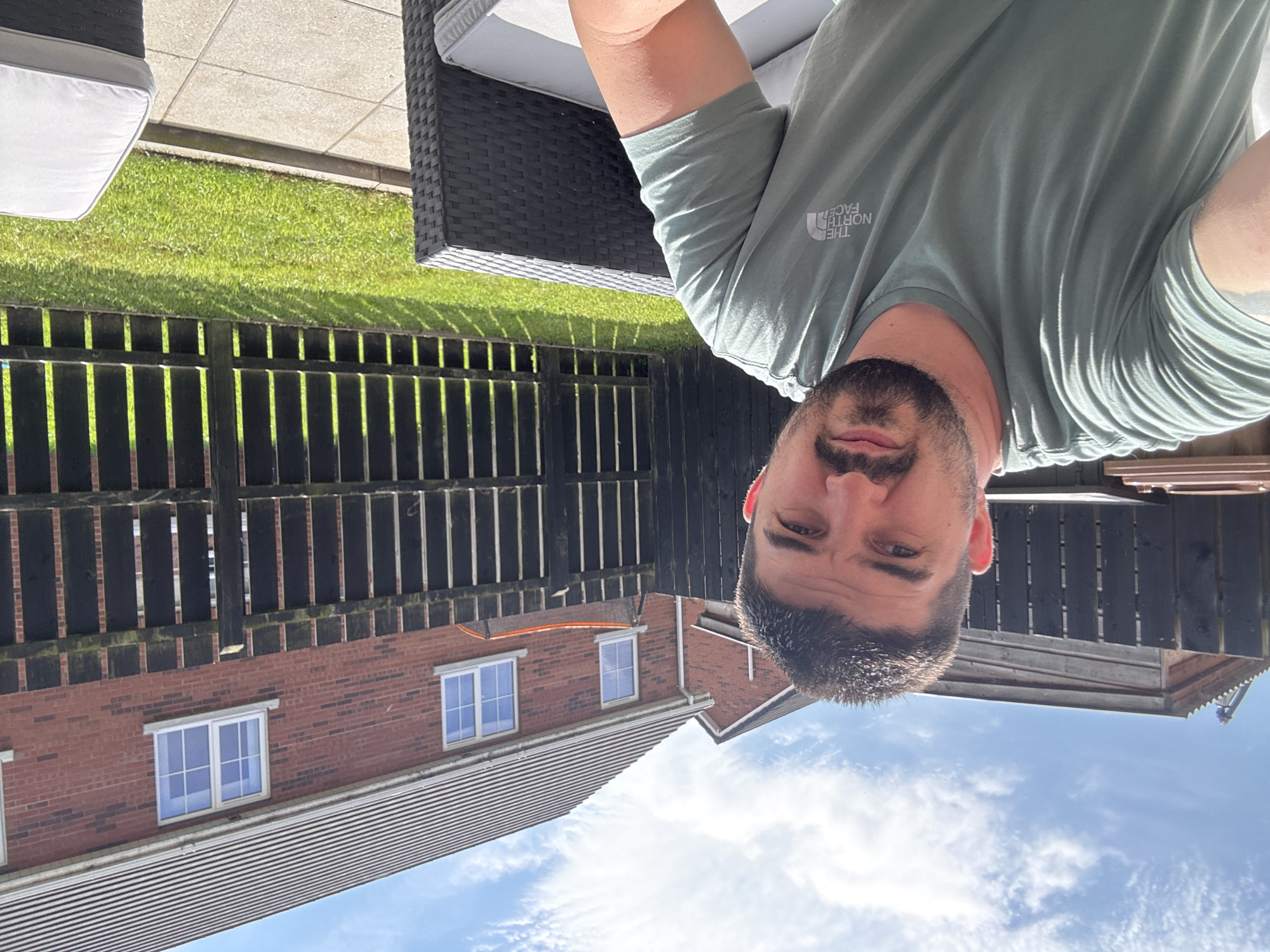 A man with dark hair and a beard is seated in a garden, taking a selfie. He is wearing a light green t-shirt with "The North Face" logo visible on the chest. The background shows a wooden fence, a well-maintained lawn, and a brick house with white-framed windows. Two pieces of outdoor furniture, a dark wicker armchair and a lighter-colored sofa, are partially visible in the foreground. The sky is blue with scattered white clouds, suggesting a sunny day in Craigavon, United Kingdom.