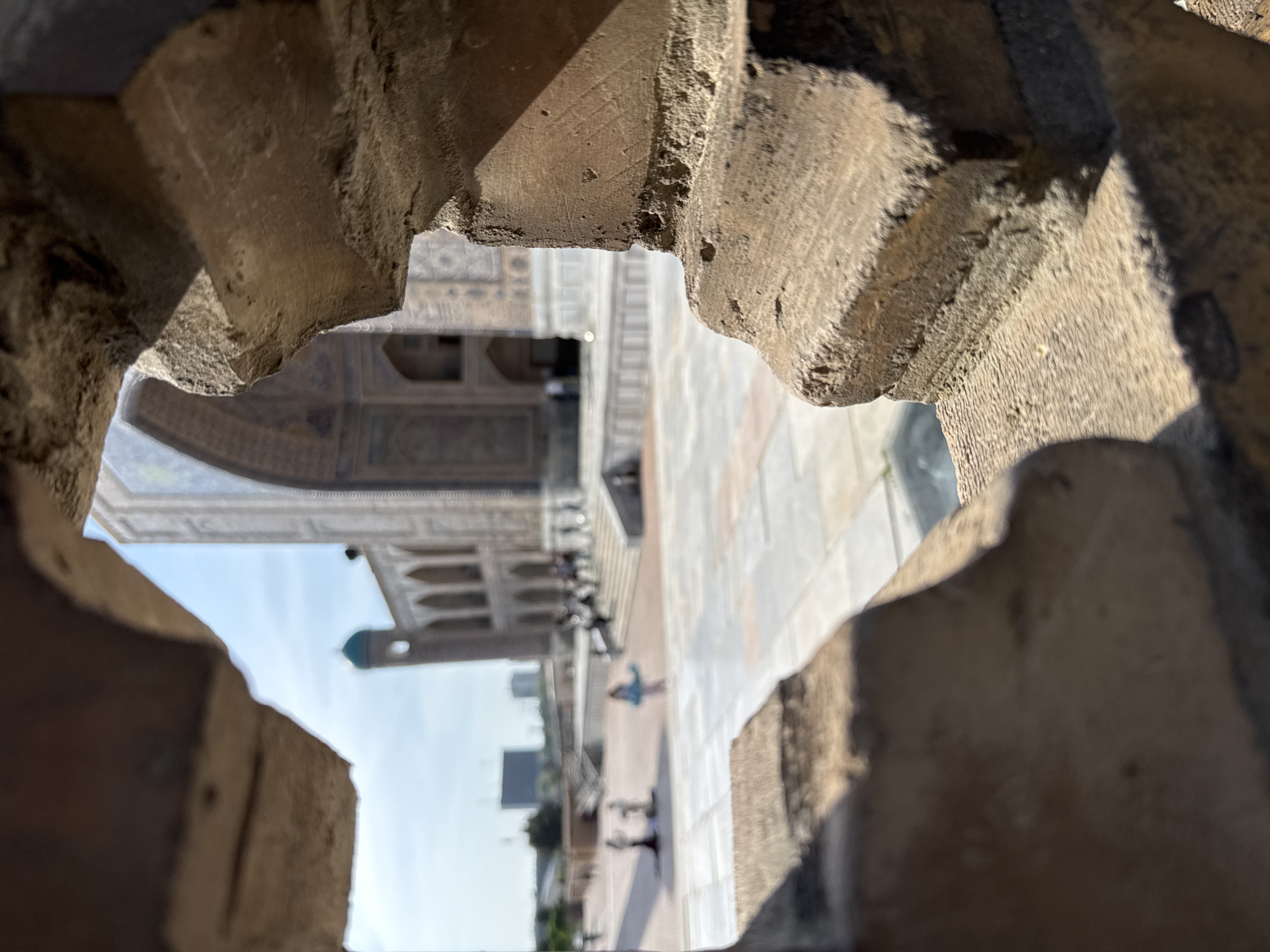 The image captures an exterior view of what appears to be the courtyard and buildings of a religious or historical site, observed through a framed stone structure. The setting is likely Samarkand City, Uzbekistan, based on the architectural style visible.
The foreground features rough-hewn, textured stone that forms an arched opening, framing the scene beyond. This stone suggests a historical structure or building element.
Through the opening, the courtyard is visible with a light-colored stone or tiled ground. Buildings surround the courtyard, exhibiting intricate tile work and arched facades, typical of Islamic architecture. A dome-like structure with a green or teal-colored top can be seen in the distance.
Several figures can be seen walking in the courtyard, suggesting the space is accessible to the public.