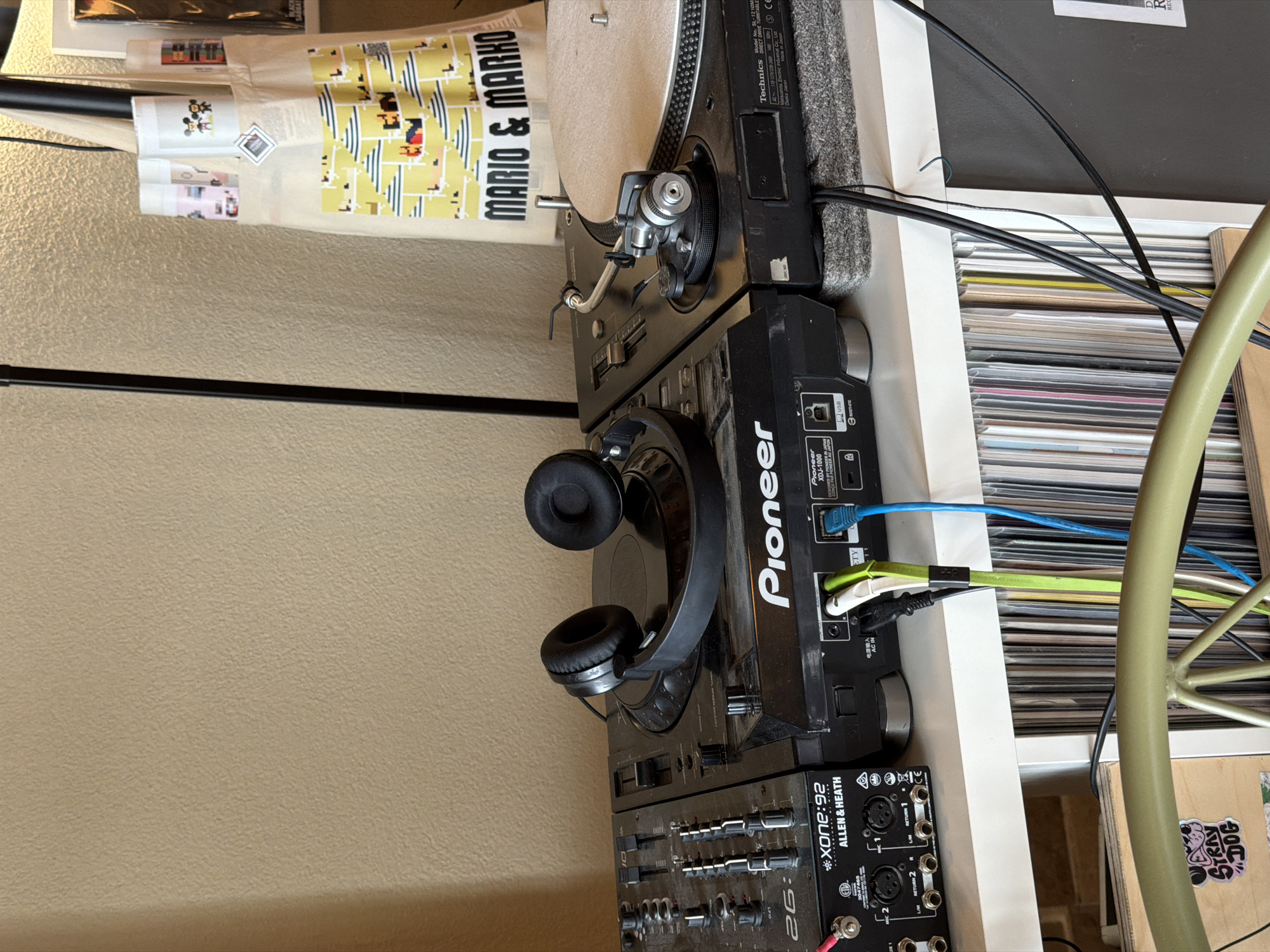 The image shows a music setup consisting of a Technics turntable, a Pioneer XDJ-1000 media player, an Allen & Heath Xone:92 mixer, and headphones. The equipment is stacked vertically on a white shelf, next to a collection of records. A tote bag with "MARIO & MARKO" printed on it is visible next to the turntable. The location of the image is unknown and not Bečići, Montenegro.